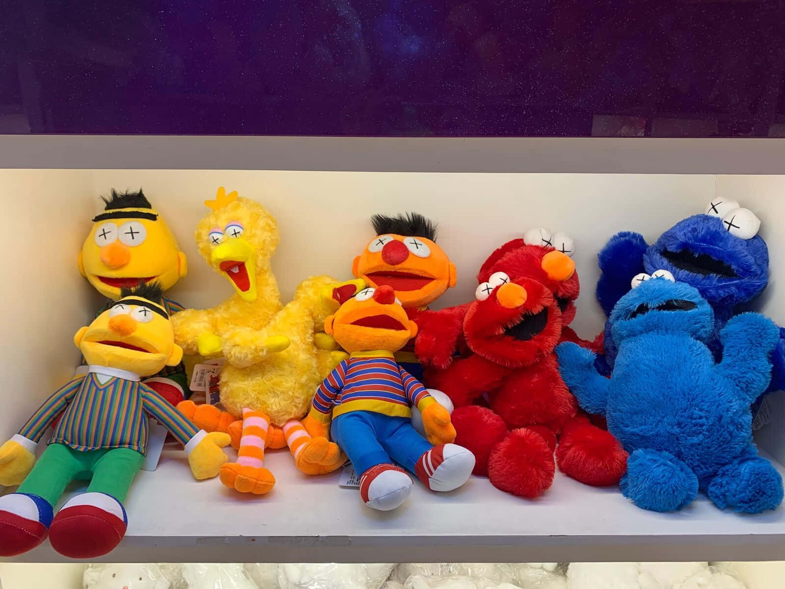 Kaws Sesame Street 桌布 壁紙 — 一排色彩繽紛、眼睛呈X形的芝麻街絨毛玩具放在白色架子上，背景是深紫色牆壁。