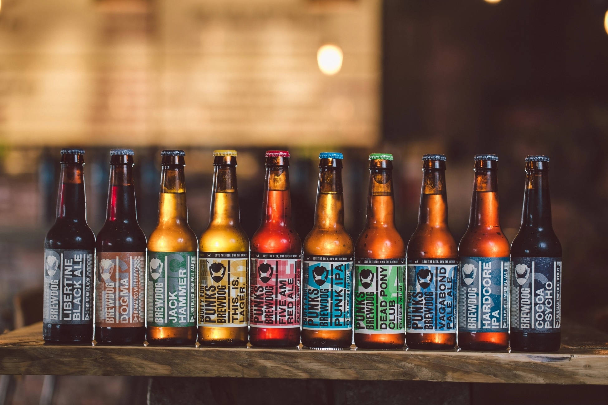 Drink 桌布 壁紙 — 一排色彩繽紛的BrewDog啤酒瓶排列在質樸的木吧上，在溫暖的環境燈光下。
