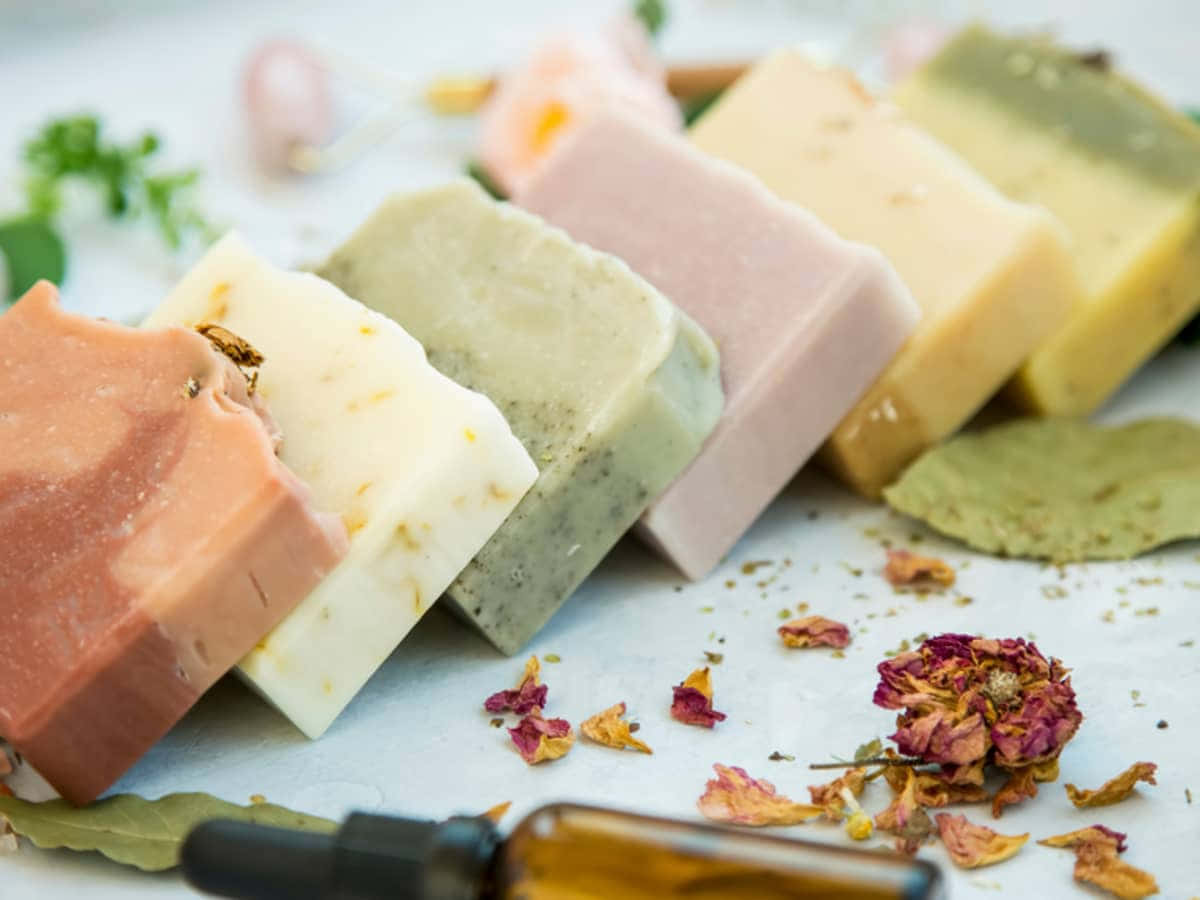 bath 桌布 壁紙 — 一排色彩繽紛的手工皂，周圍散落著乾花瓣和香草，置於白色表面上。