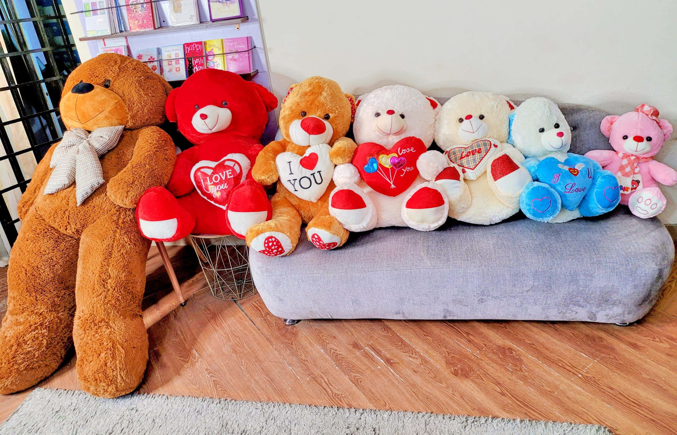 Teddy Bear 桌布 壁紙 — 一排色彩繽紛的毛絨泰迪熊帶有心形主題訊息，坐在灰色沙發上，處於一個舒適的室內環境。