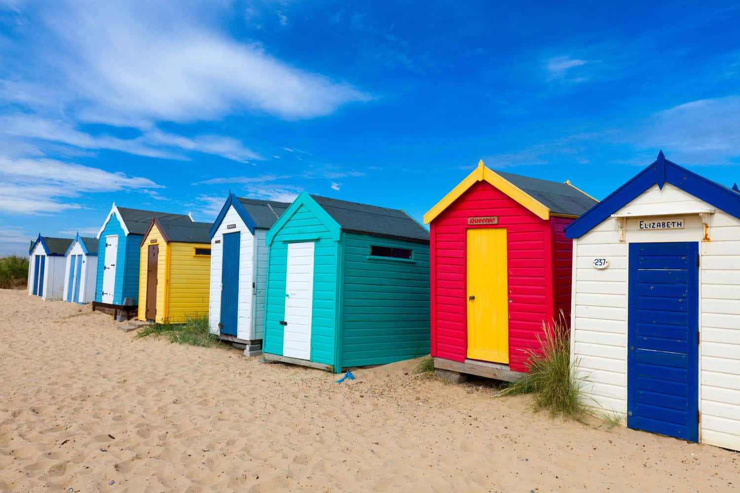 Beach Huts 桌布 壁紙 — 一排色彩繽紛的沙灘小屋整齊排列在金色沙灘上，上方是明亮藍天與薄雲。