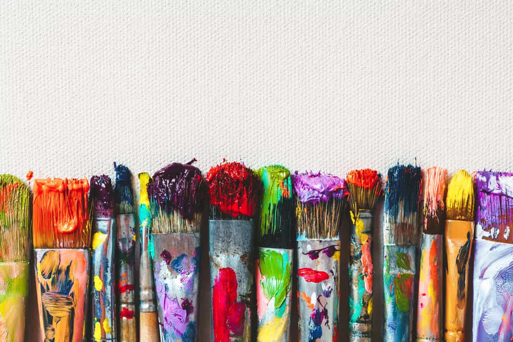 Paint Brush 桌布 壁紙 — 一排色彩繽紛的畫筆，帶有鮮豔的乾燥顏料塊，靠在有紋理的白色畫布背景上。