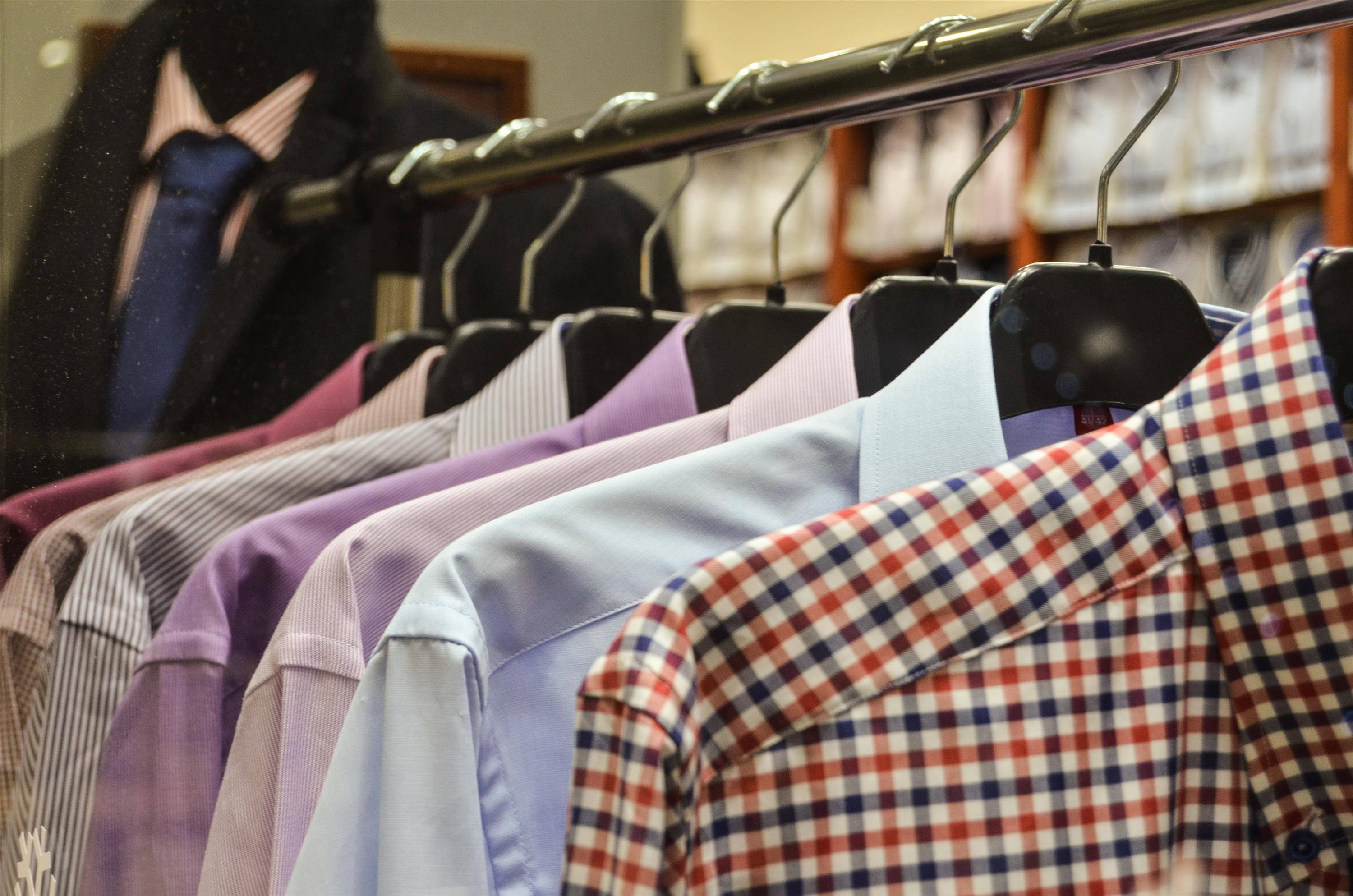 Clothes 桌布 壁紙 — 一排色彩繽紛的襯衫整齊地掛在黑色衣架上，位於光線充足的男裝店內，背景有一個西裝模特兒。