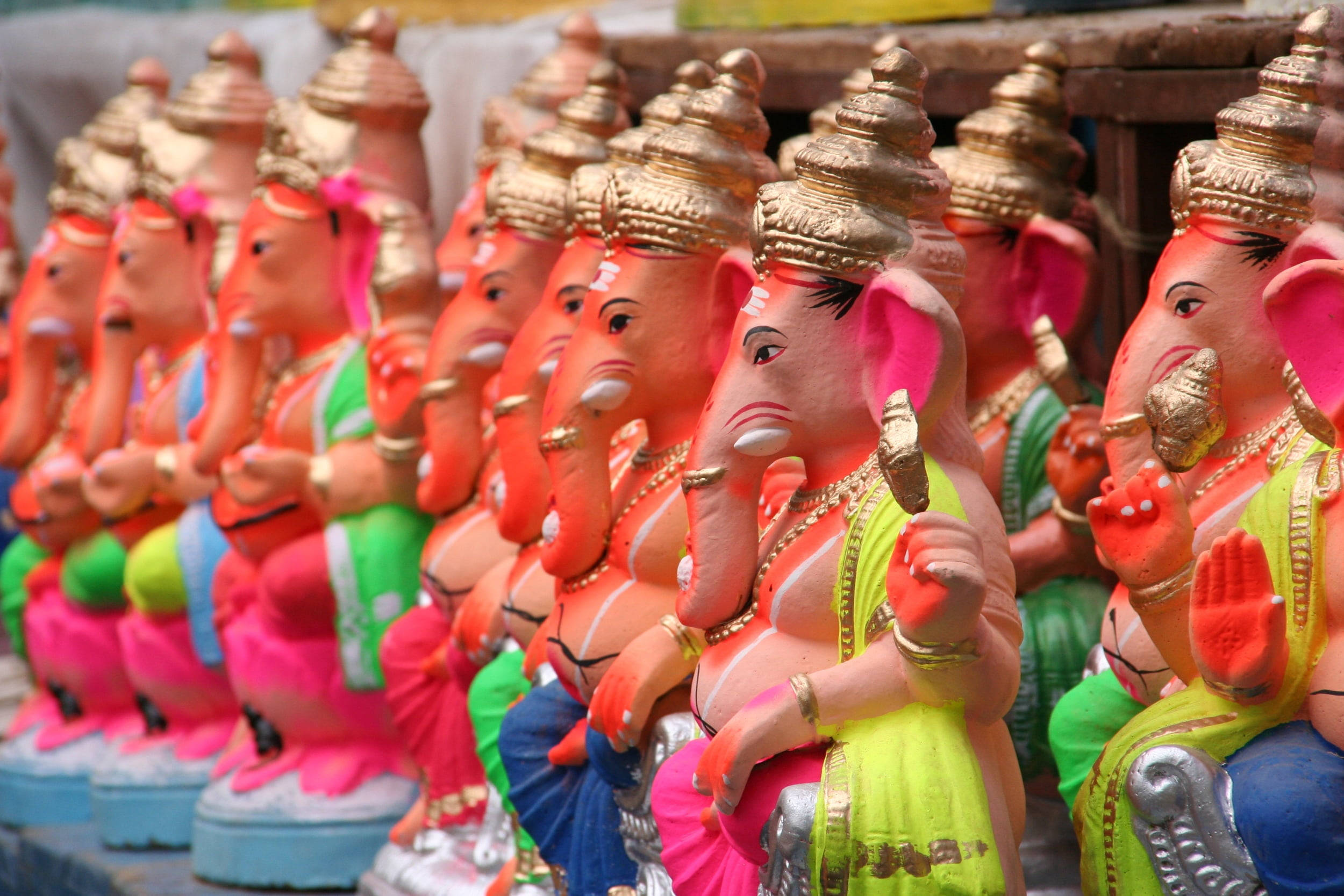 Ganesh 桌布 壁紙 — 一排色彩繽紛的象神雕像，身穿鮮豔的粉紅色、綠色和金色服飾，整齊排列出售。