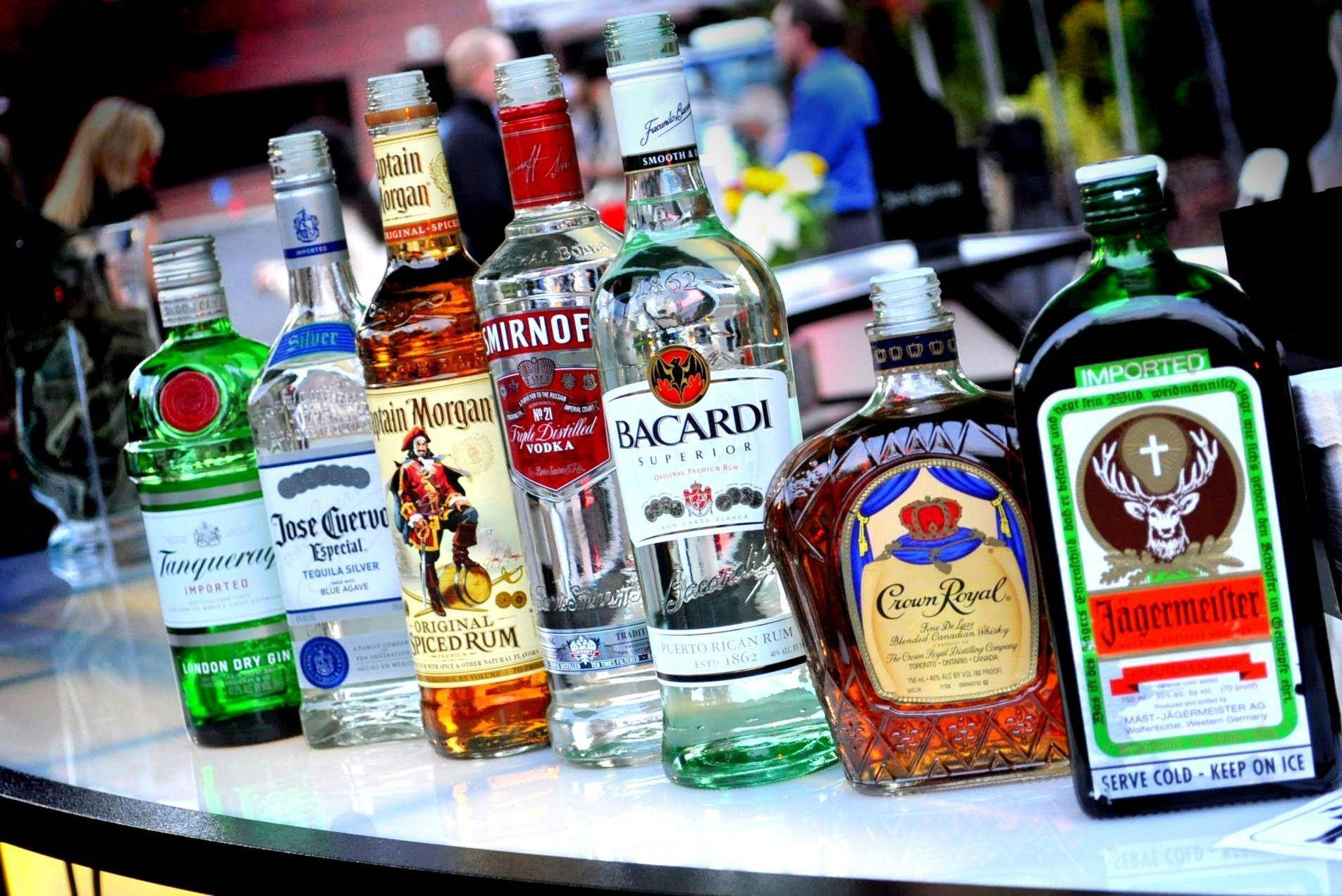 alcohol 桌布 壁紙 — 一排色彩繽紛的酒瓶，包括Jagermeister、Bacardi和Crown Royal，放在吧檯上，背景有模糊的顧客。