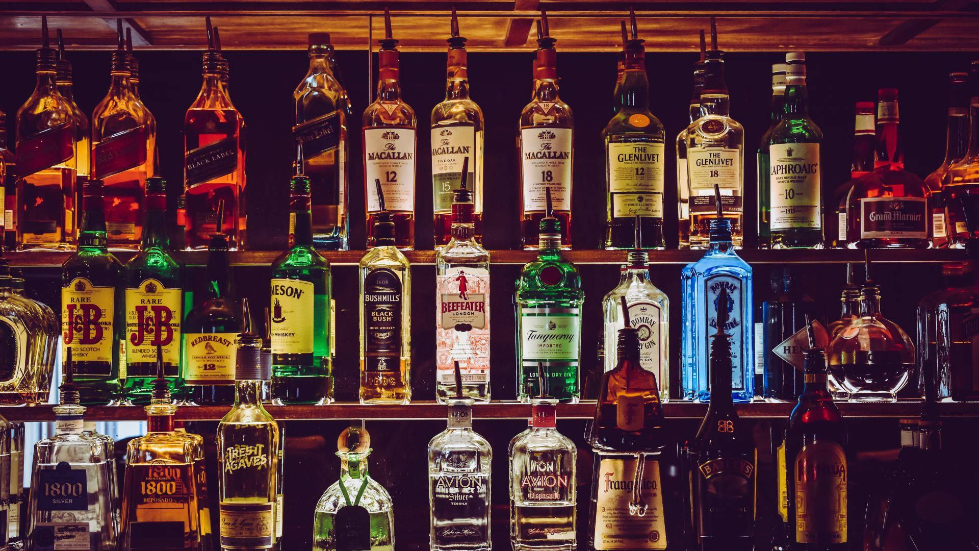 drinks 桌布 壁紙 — 一排色彩繽紛的酒瓶在溫暖的環境燈光照射下，排列在黑暗的酒吧架上。