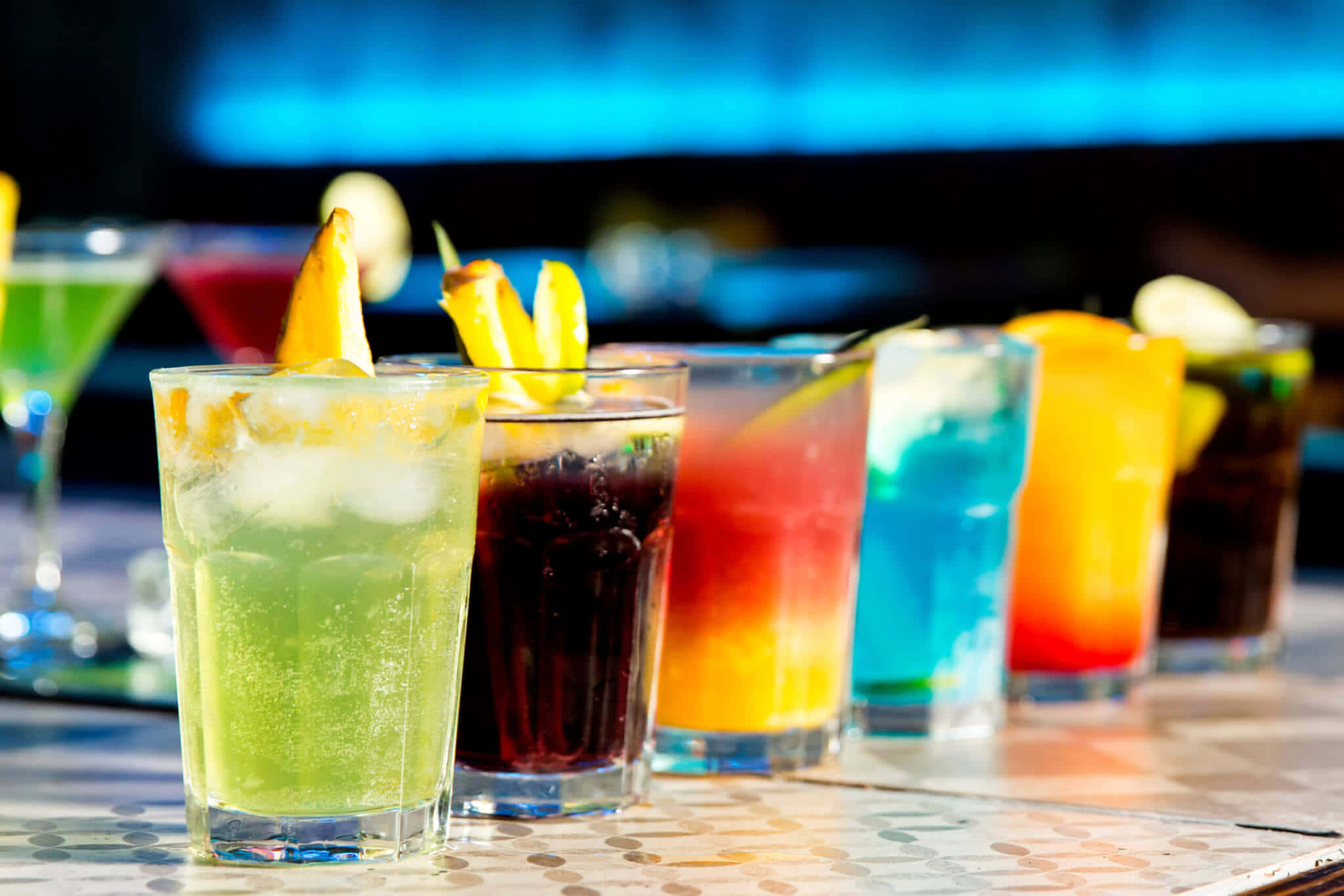 Beautiful Drink 桌布 壁紙 — 一排色彩繽紛的雞尾酒配上柑橘裝飾，放在有圖案的吧檯上，在明亮的燈光下。
