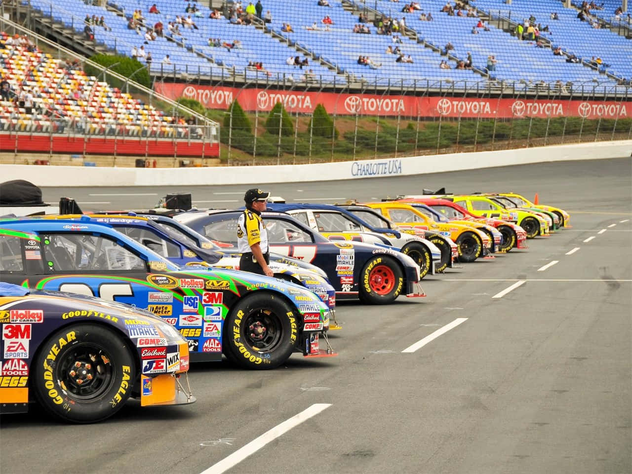 美國 桌布 壁紙 — 一排色彩鮮豔的NASCAR賽車整齊排列在賽道上，旁邊站著一名賽道工作人員，天空部分多雲。