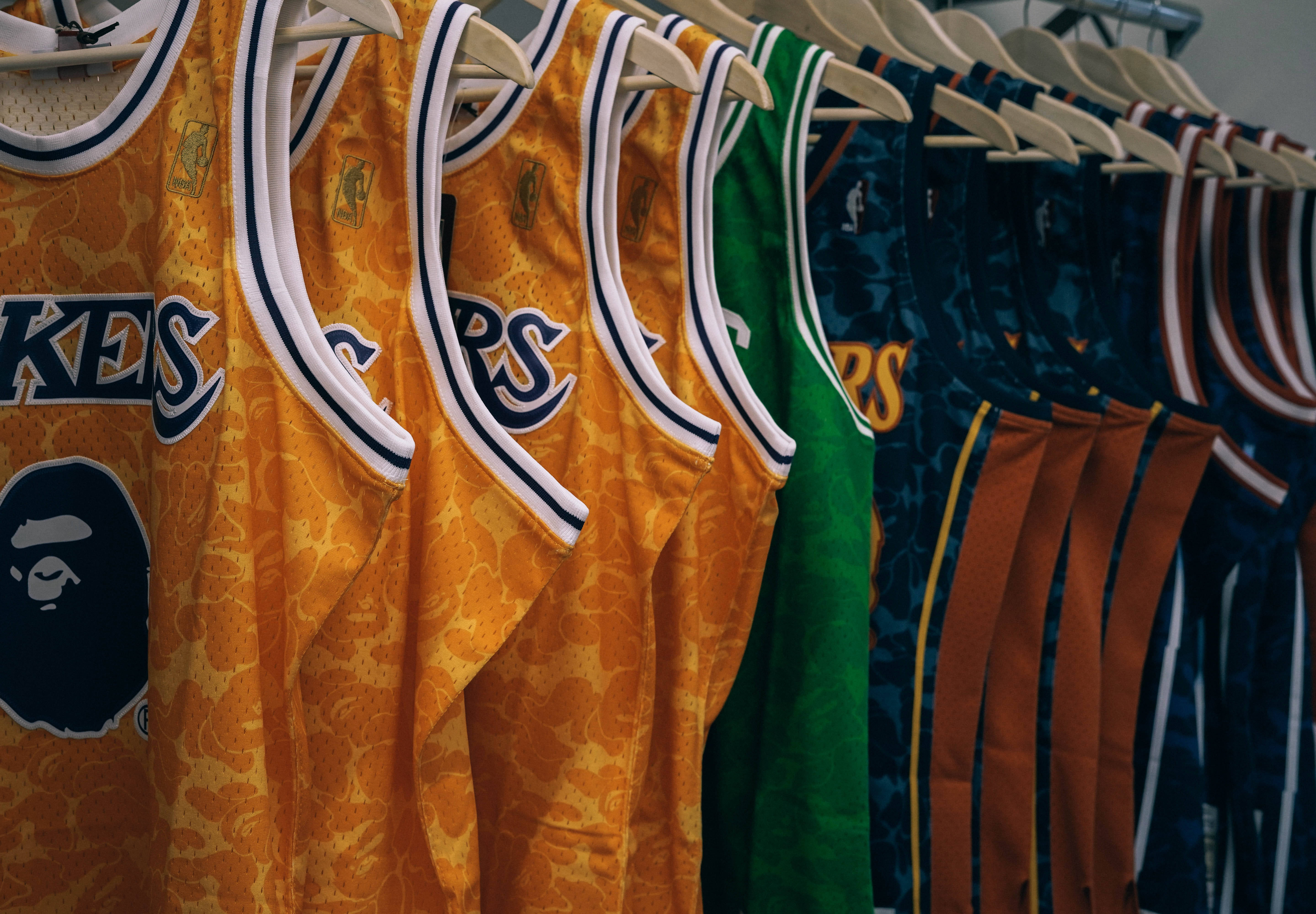 Clothes 桌布 壁紙 — 一排色彩鮮豔的NBA球衣，帶有迷彩圖案和BAPE標誌，整齊地掛在零售展示的木衣架上。