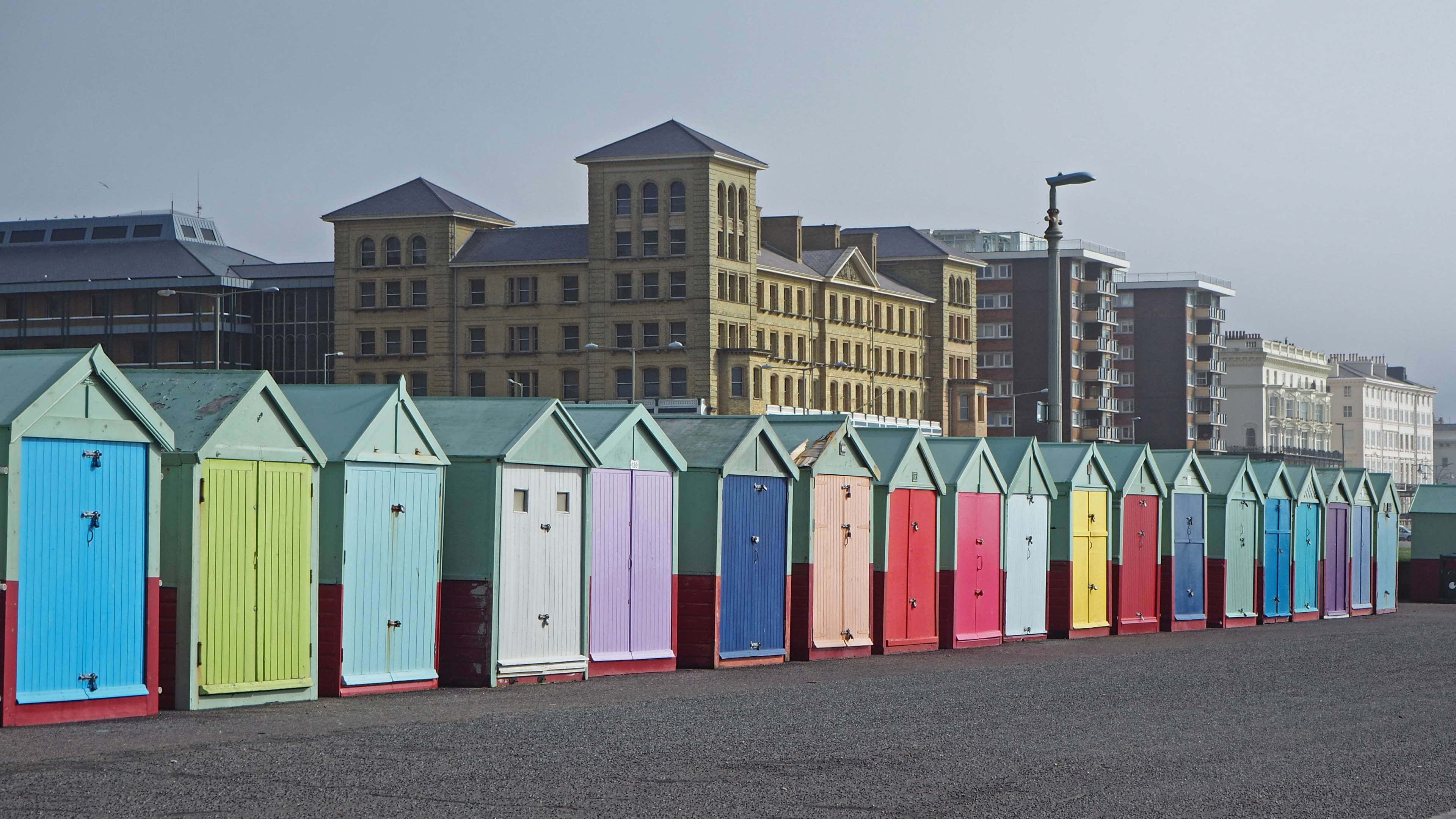 Beach Huts 桌布 壁紙 — 一排色彩鮮豔的沙灘小屋排列在鋪砌的海濱長廊上，背景是歷史建築，天空多雲。