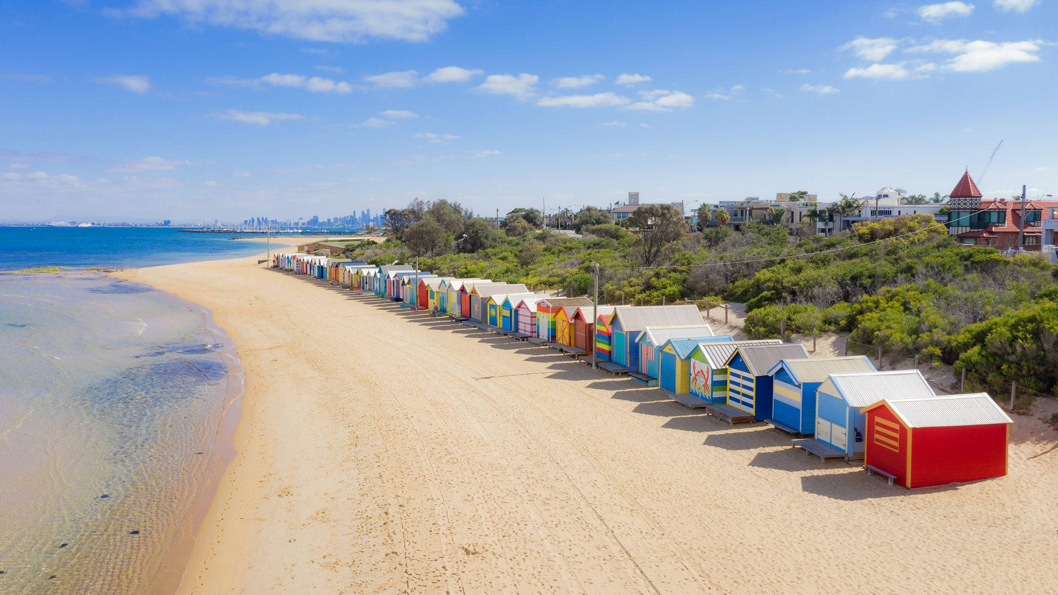 Melbourne 桌布 壁紙 — 一排色彩鮮豔的沙灘小屋沿著金色沙灘排列，在清澈的藍天下，遠處有城市天際線。