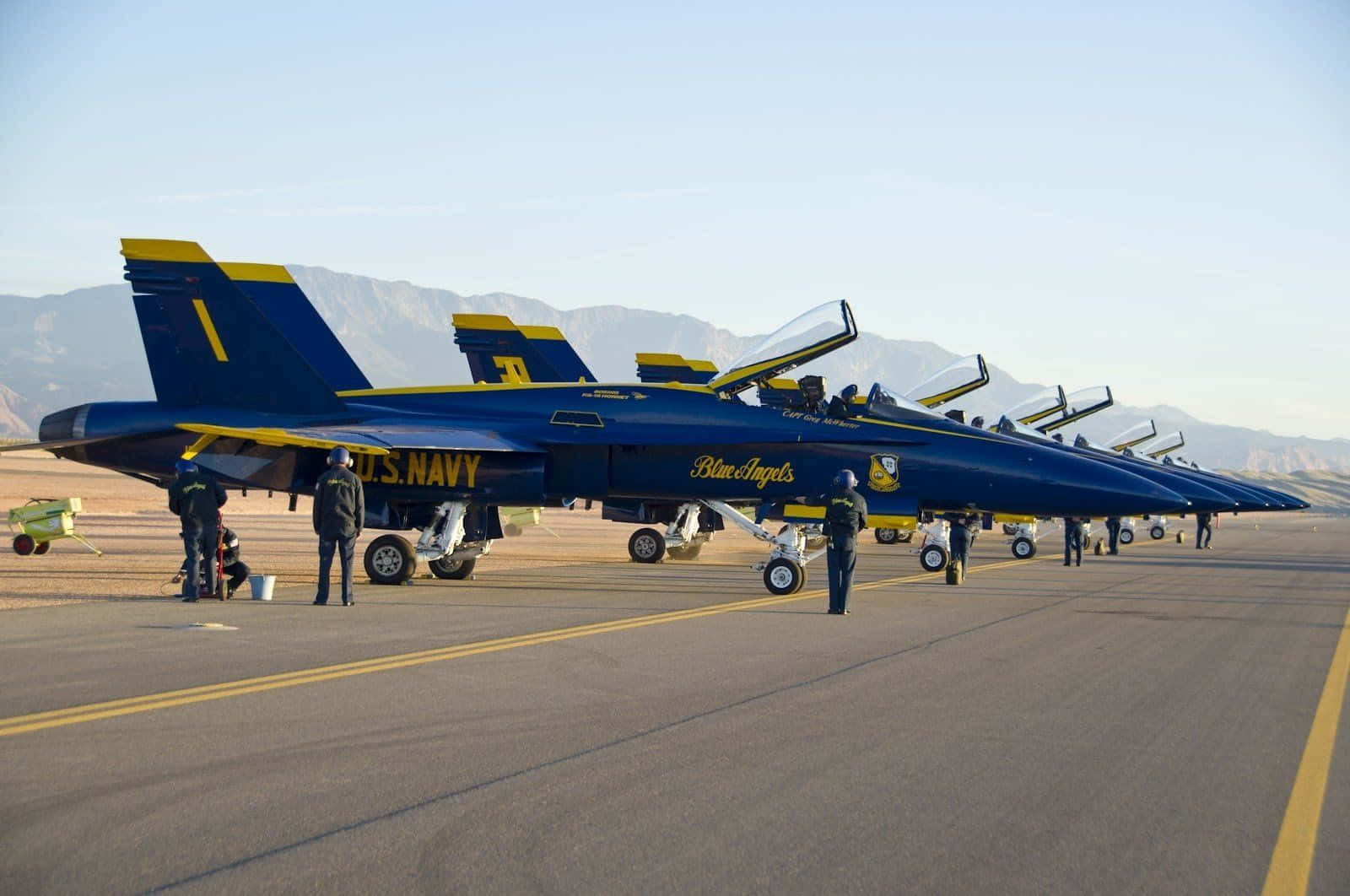 US Navy 桌布 壁紙 — 一排藍天使戰鬥機停在機場上，機組人員在晴朗的天空下，以遠處的山脈為背景，照顧著它們。