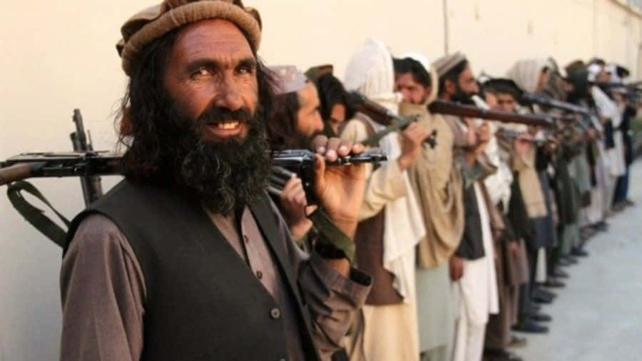Taliban 桌布 壁紙 — 一排身穿傳統服飾的武裝男子在明亮的日光下，對著素色牆壁列隊站立。