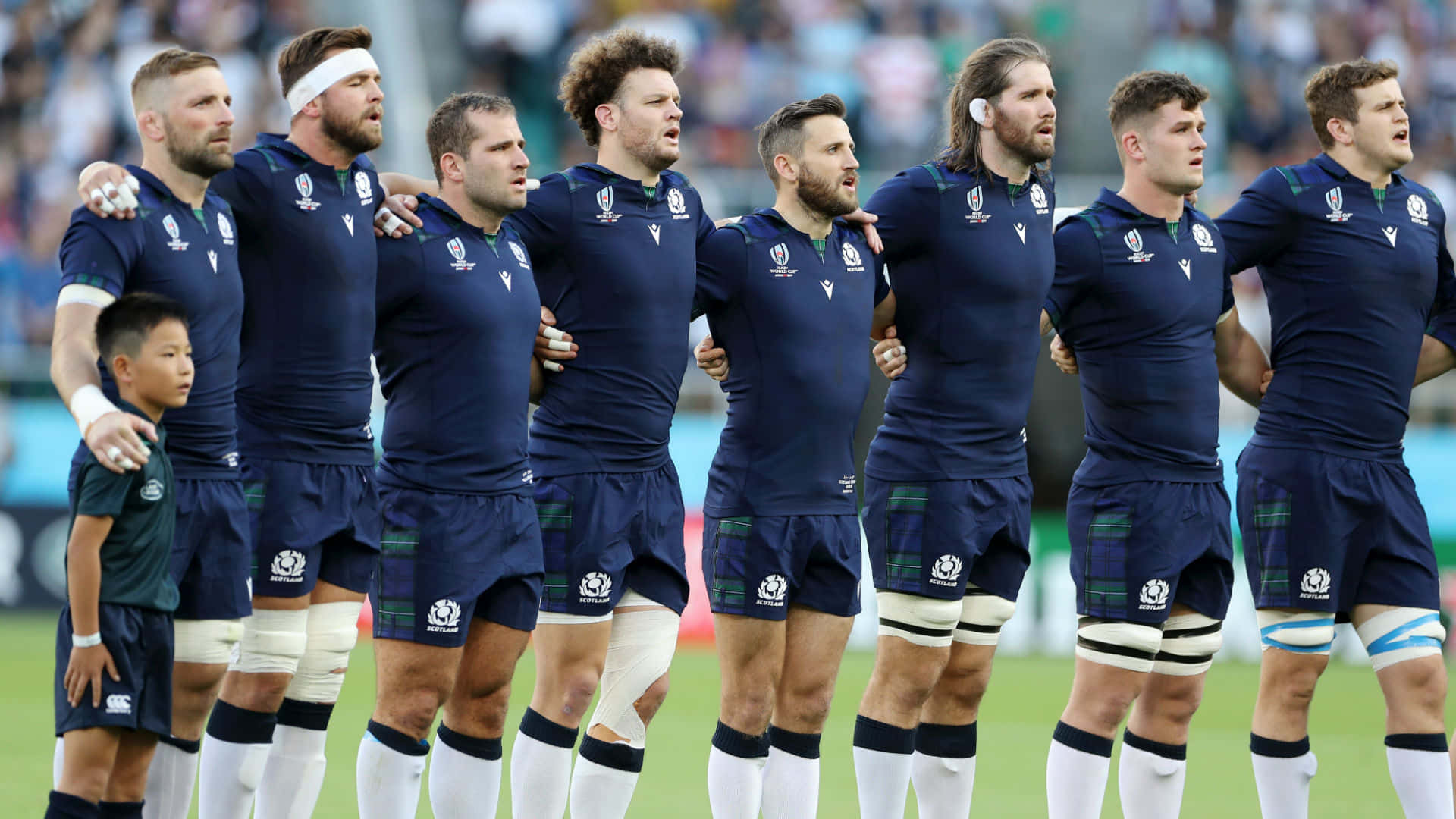 Scotland Rugby 桌布 壁紙 — 一排身穿海軍藍球衣的蘇格蘭橄欖球運動員在國歌奏起時，在綠色球場上莊嚴地互相搭肩站立。