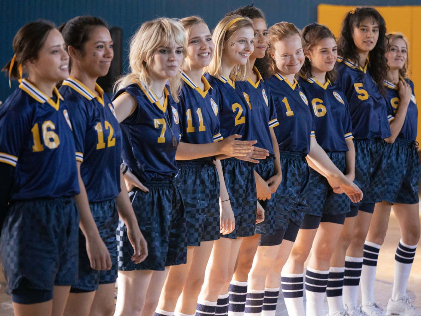 Cheerleader 桌布 壁紙 — 一排身穿藍黃色制服的年輕女排球員自信地站在球場上，微笑著擺姿勢給相機拍照。