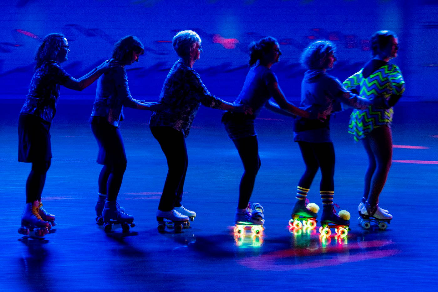 skating 桌布 壁紙 — 一排輪滑者在霓虹燈下滑行，發光的輪子在黑暗的溜冰場上反射。