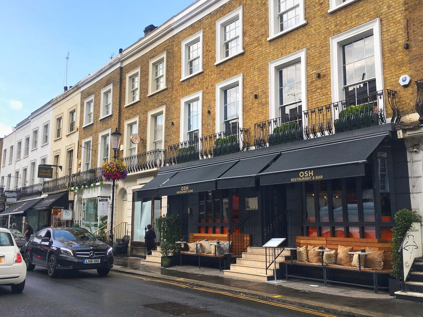 Restaurant 桌布 壁紙 — 一排迷人的磚砌聯排別墅，帶有黑色遮陽篷和戶外座位，排列在一條潮濕的城市街道上，天空清澈湛藍。