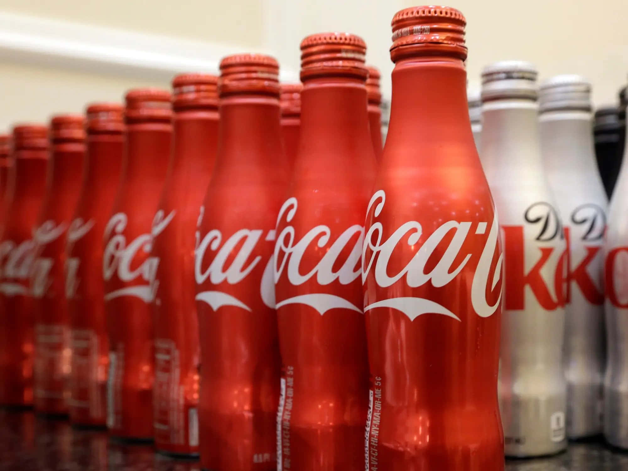 coca cola 桌布 壁紙 — 一排閃亮的紅色可口可樂瓶，帶有白色草書標誌，與銀色健怡可樂瓶在深色檯面上清晰對焦。