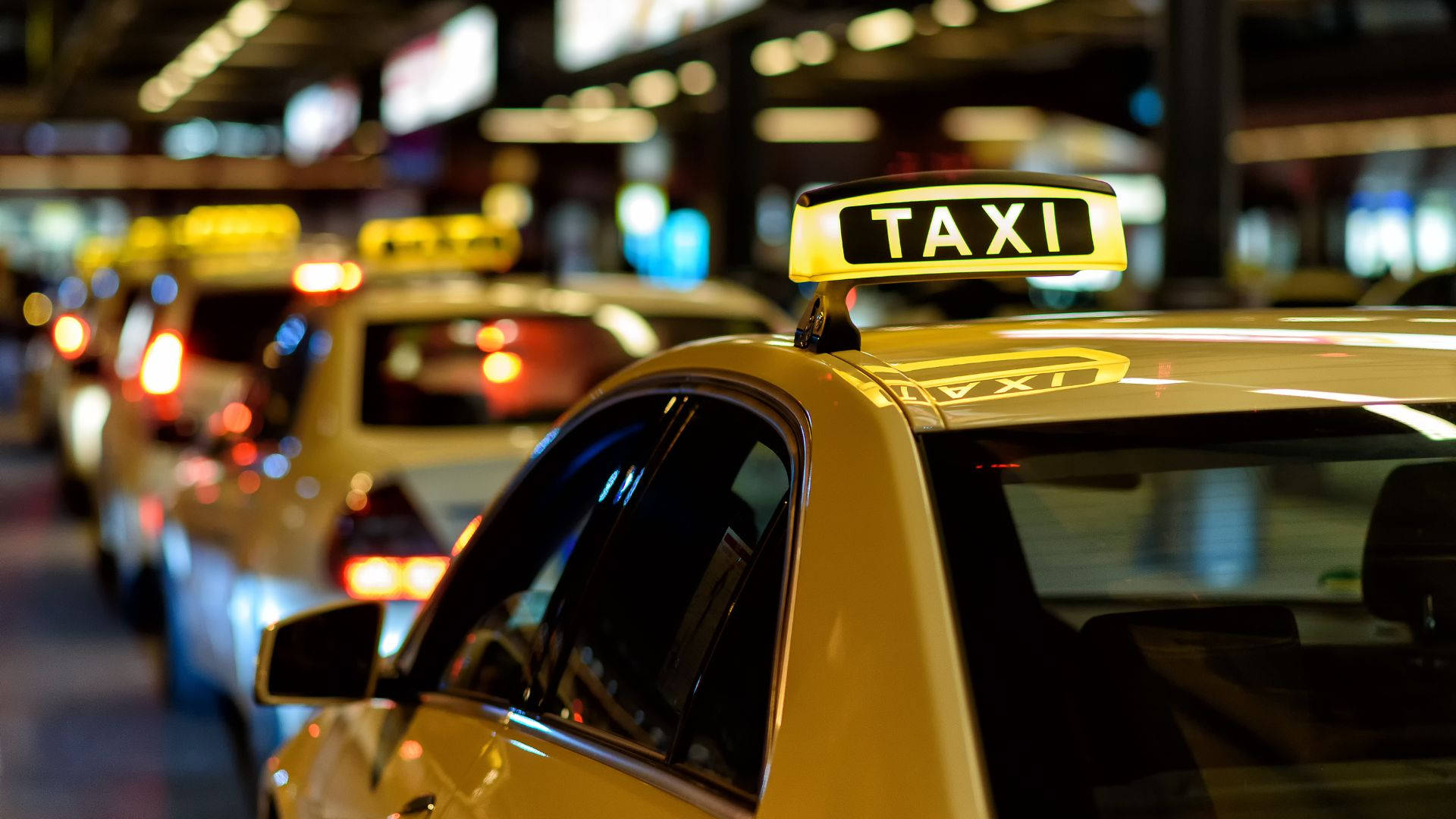 taxi 桌布 壁紙 — 一排黃色的士在夜晚排列，帶有發光的「TAXI」標誌和模糊的城市燈光作為背景。