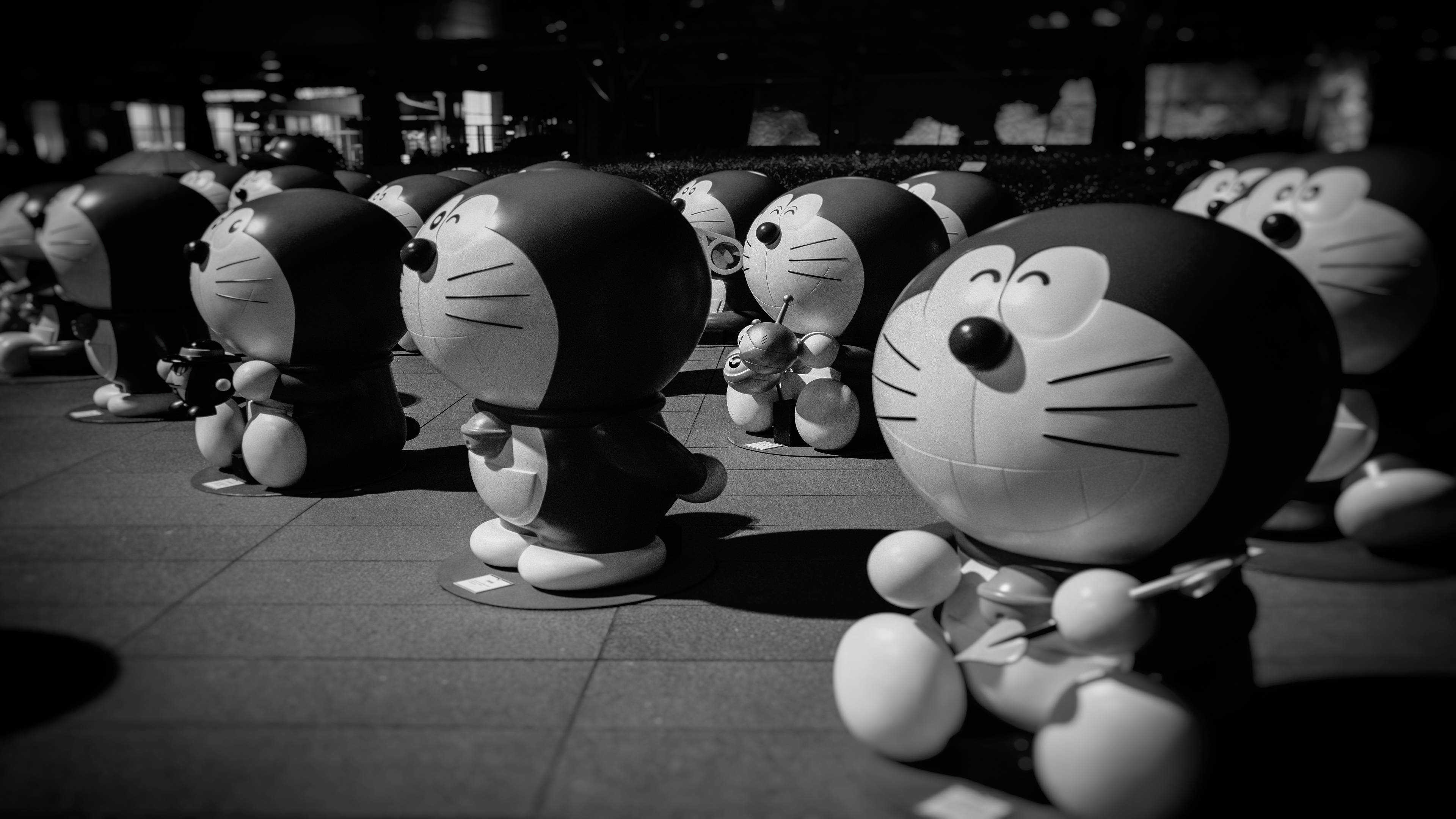 Doraemon 桌布 壁紙 — 一排黑白哆啦A夢雕像站在瓷磚廣場上，夜間燈光投射出戲劇性的陰影。