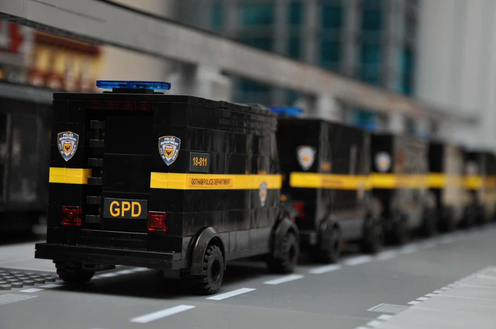 Gotham City Police Department 桌布 壁紙 — 一排黑色 LEGO 高譚市警察局廂型車，帶有黃色條紋和藍色燈光，停放在城市街道上。