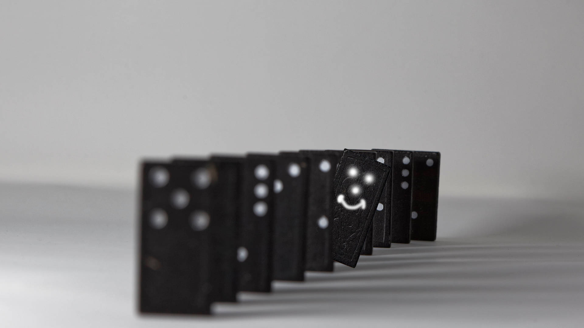 dominos 桌布 壁紙 — 一排黑色骨牌，其中一個笑臉牌在柔和的灰色背景上傾倒。