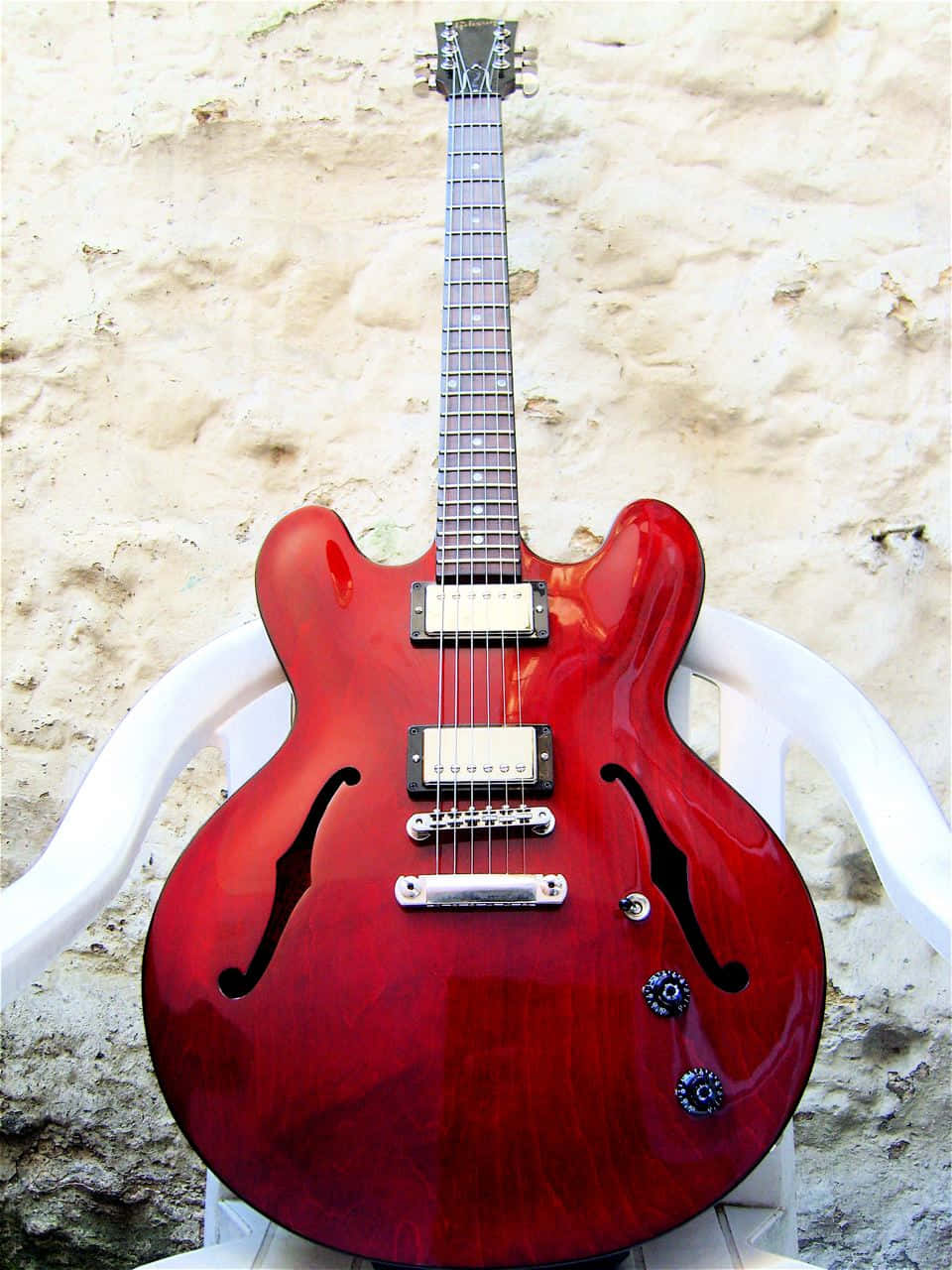 Guitar Aesthetic 桌布 壁紙 — 一支亮面紅色半空心電吉他直立放在白色塑膠椅上，背景是質感豐富的米色石牆。