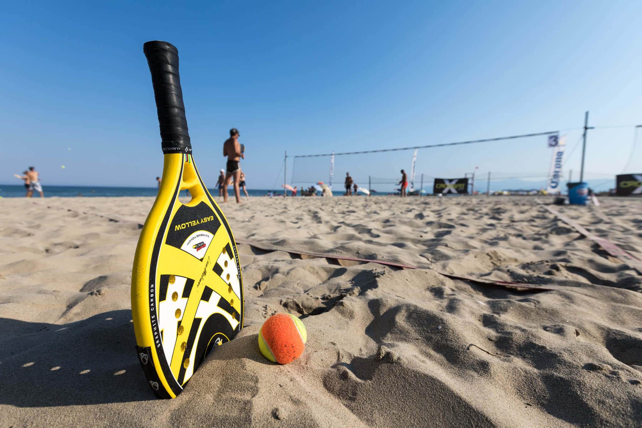 Beach Tennis 桌布 壁紙 — 一支亮黃色沙灘網球拍和橙色球放在陽光照耀的沙灘上，背景有球員和球網。