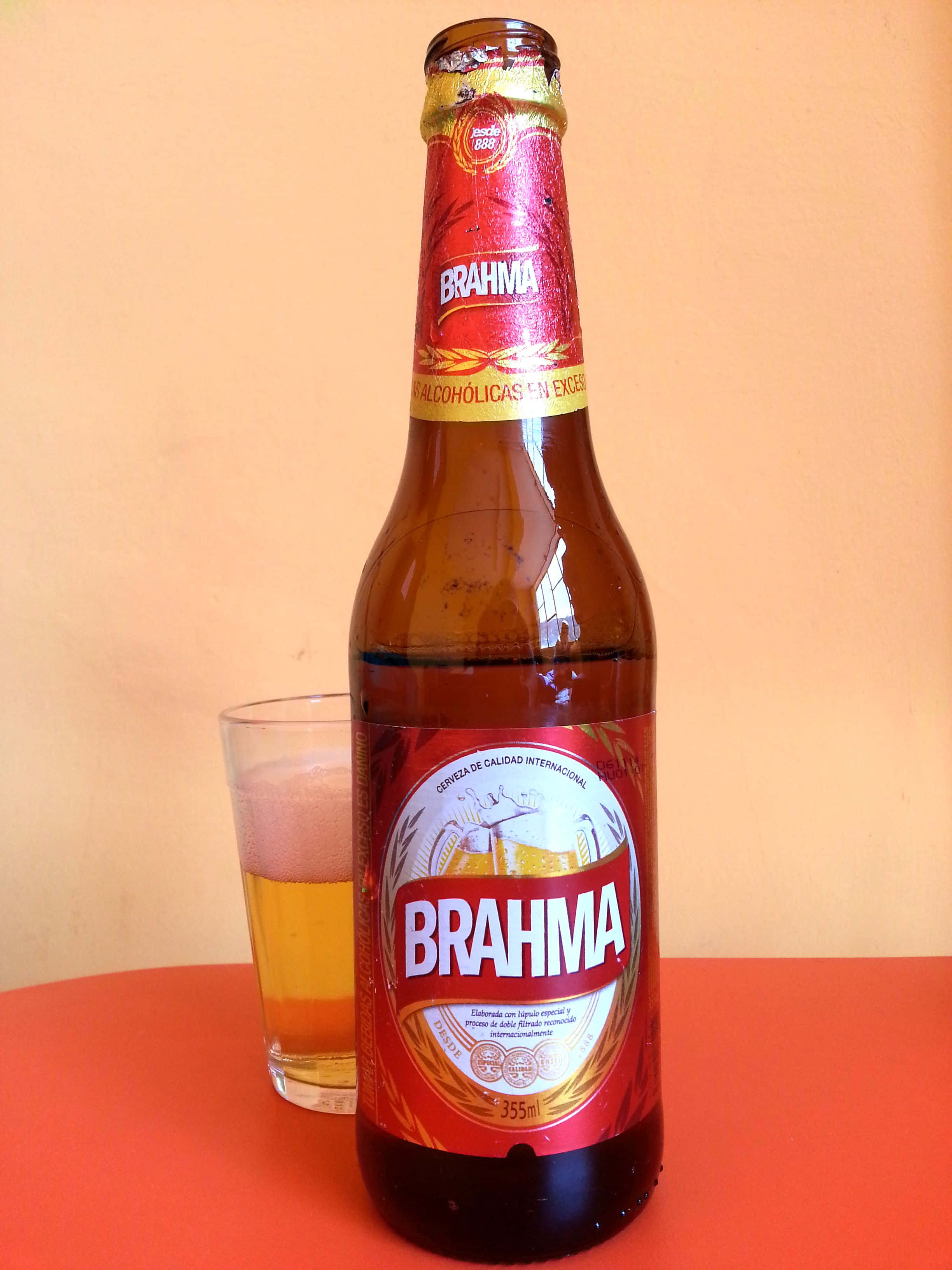 alcohol 桌布 壁紙 — 一支冰涼的Brahma啤酒瓶和旁邊一個裝滿的玻璃杯，放在鮮紅色的表面上，背景是溫暖的桃色牆壁。