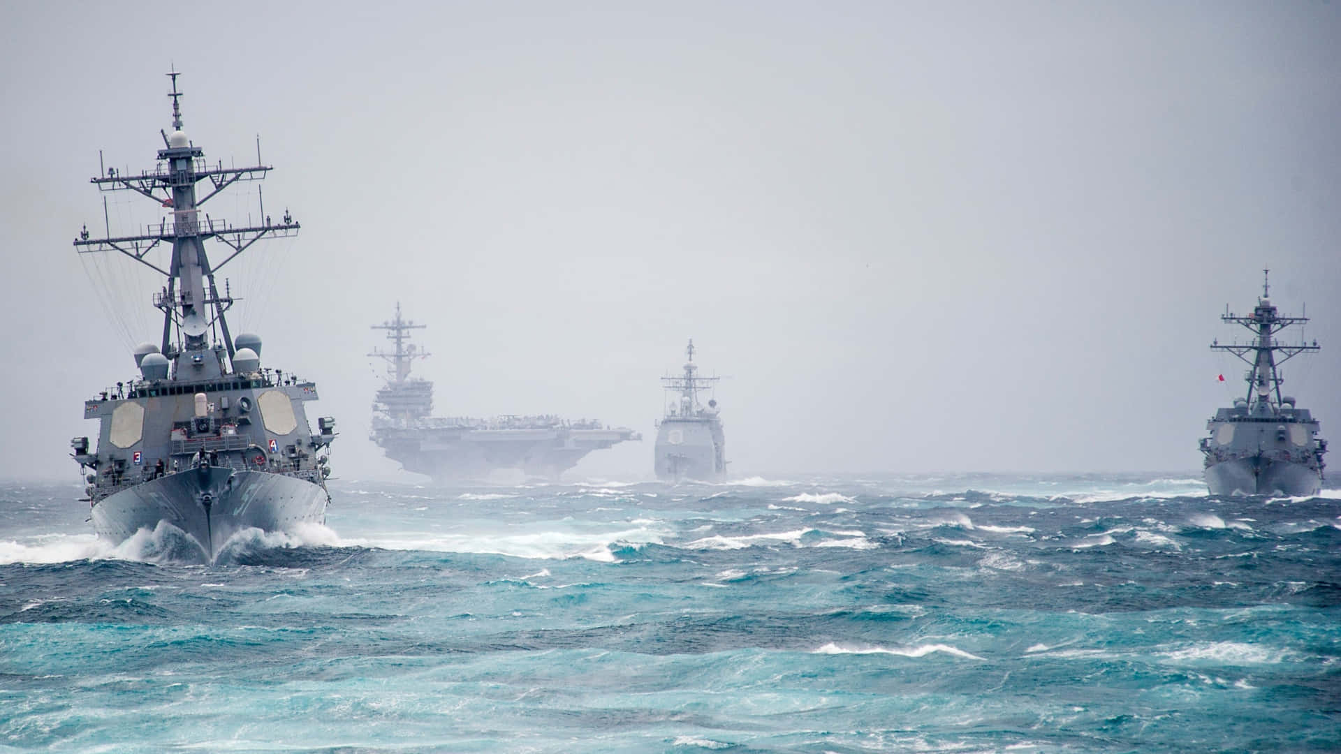Military 桌布 壁紙 — 一支包括航空母艦在內的軍艦艦隊，在多雲的天空下航行於波濤洶湧的碧綠海域。