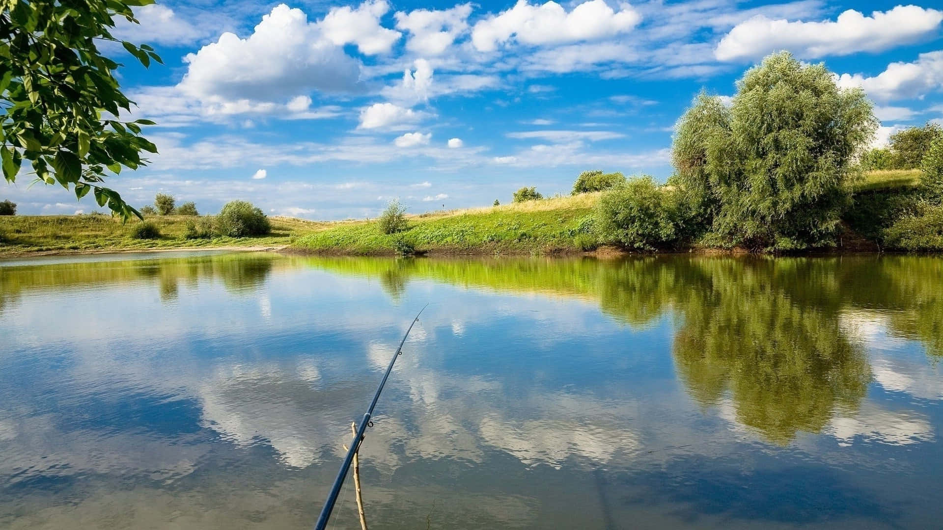 Fishing Rod 桌布 壁紙 — 一支寧靜的釣竿伸展在平靜的河面上，倒映著明亮的藍天、蓬鬆的白雲和茂密的綠樹。