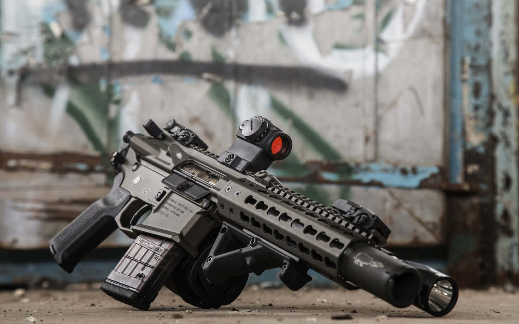 Rifle 桌布 壁紙 — 一支帶有紅點瞄準器的黑色戰術步槍放在佈滿塗鴉的牆前的碎石地上。