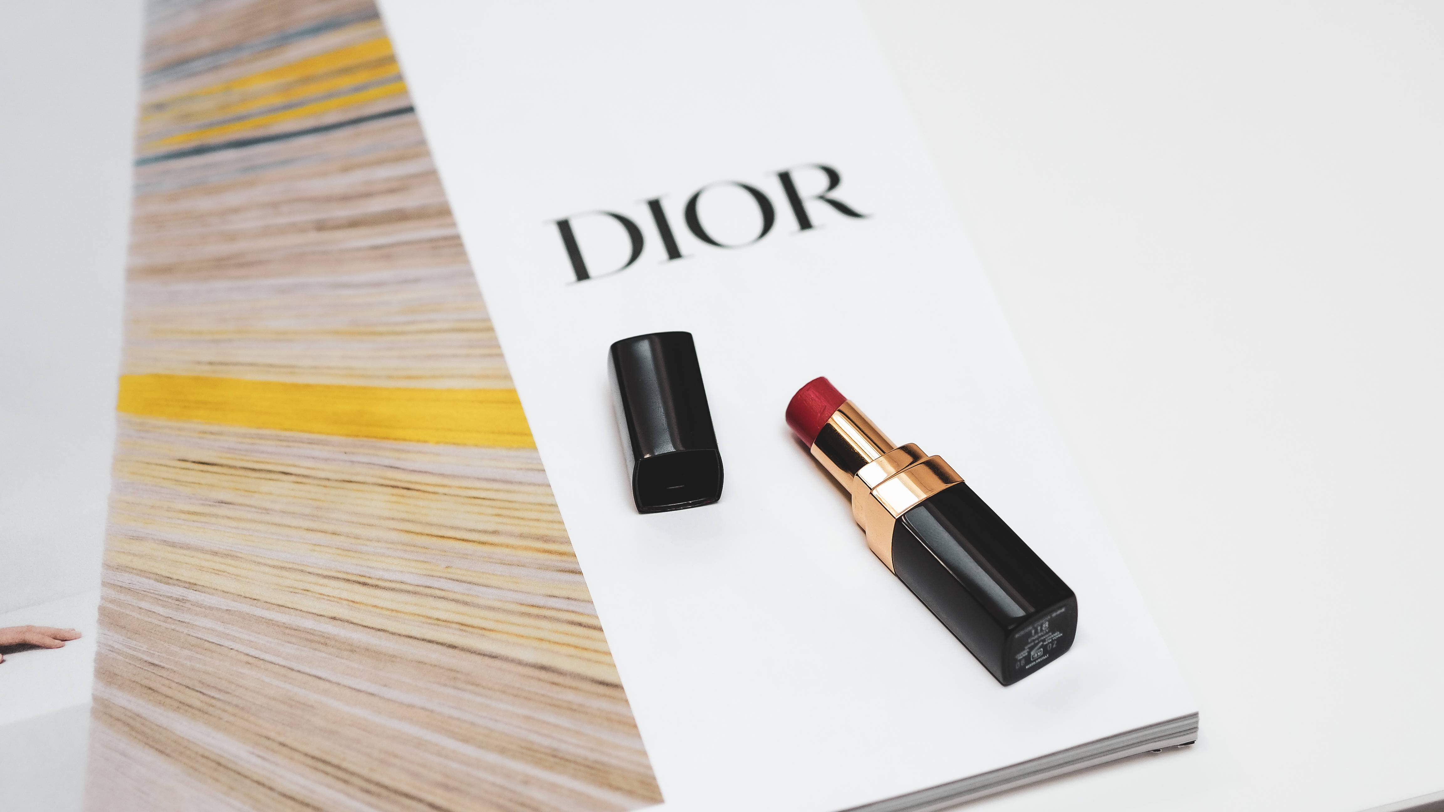 Dior 動態 桌布 壁紙 — 一支帶有金色裝飾的Dior唇膏打開放在其蓋子旁邊，位於一本白色雜誌頁面上，頁面顯示品牌名稱和一張金髮特寫。