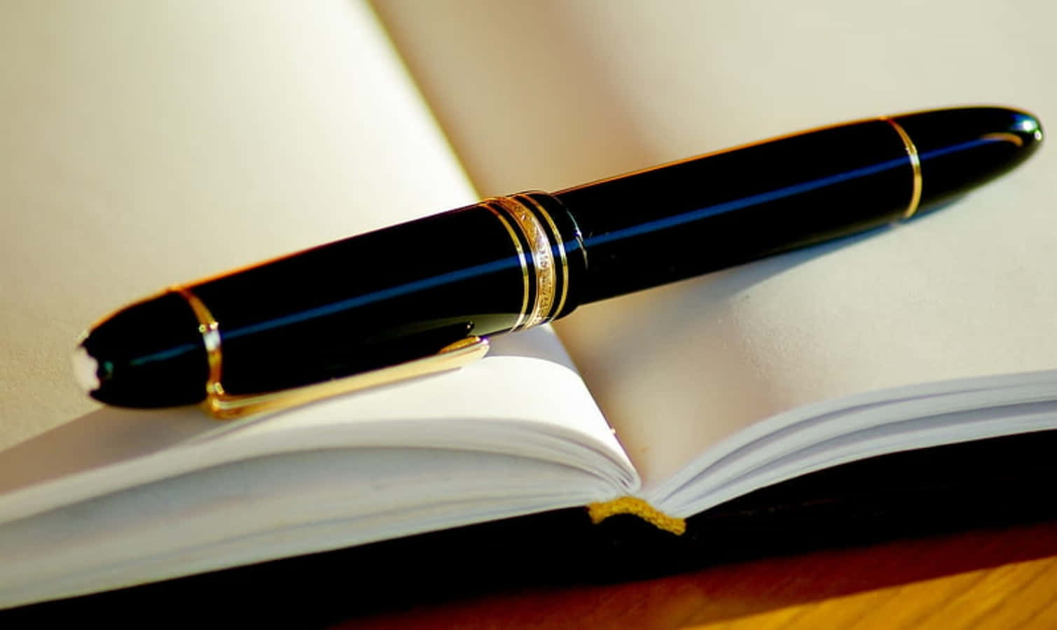 Pen 桌布 壁紙 — 一支帶有金色飾邊的時尚黑色鋼筆，斜放在空白筆記本的打開頁面上。