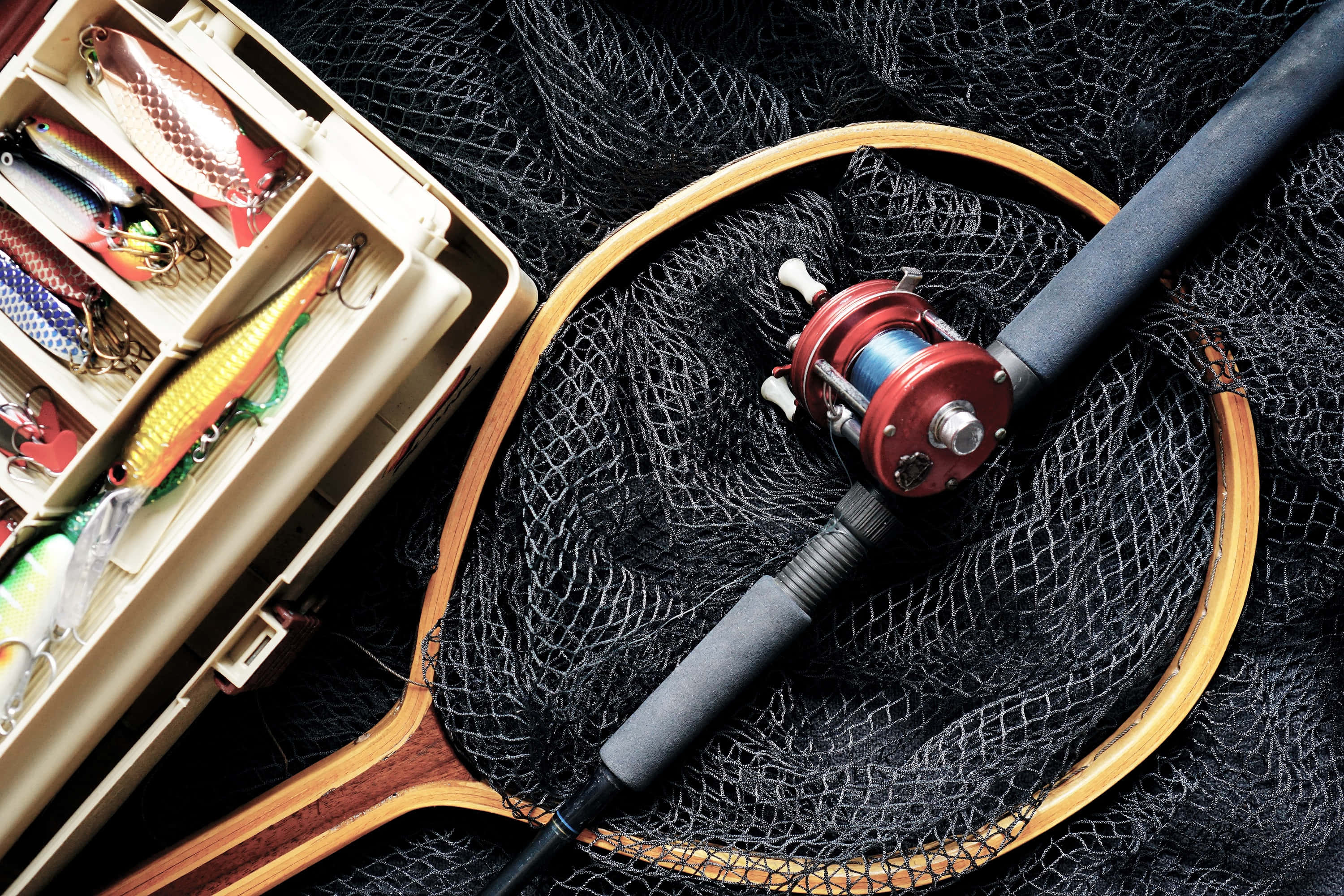 Fishing Rod 桌布 壁紙 — 一支帶紅色捲線器的釣魚竿擱在黑色網上，旁邊是一個裝滿彩色假餌的釣具箱。