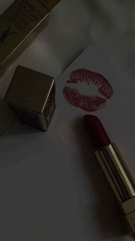 Kiss 桌布 壁紙 — 一支帶金色外殼的濃郁酒紅色唇膏，旁邊放著它的盒子和在昏暗燈光下紙上大膽的紅色唇印。