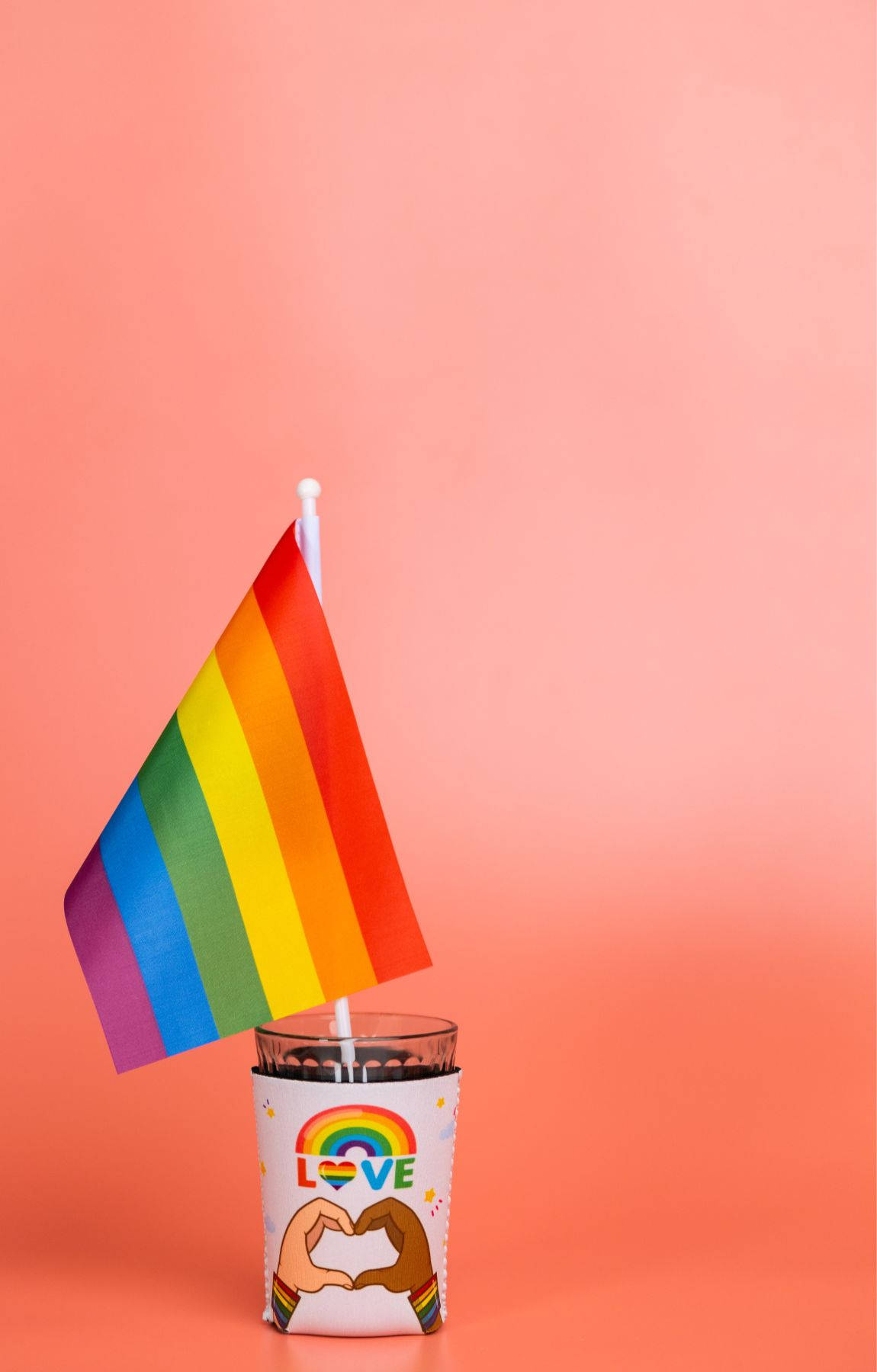 Subtle Lgbt 桌布 壁紙 — 一支彩虹旗從一個帶有彩虹心形和兩隻手形成心形符號的杯子中伸出，背景為純色蜜桃色。