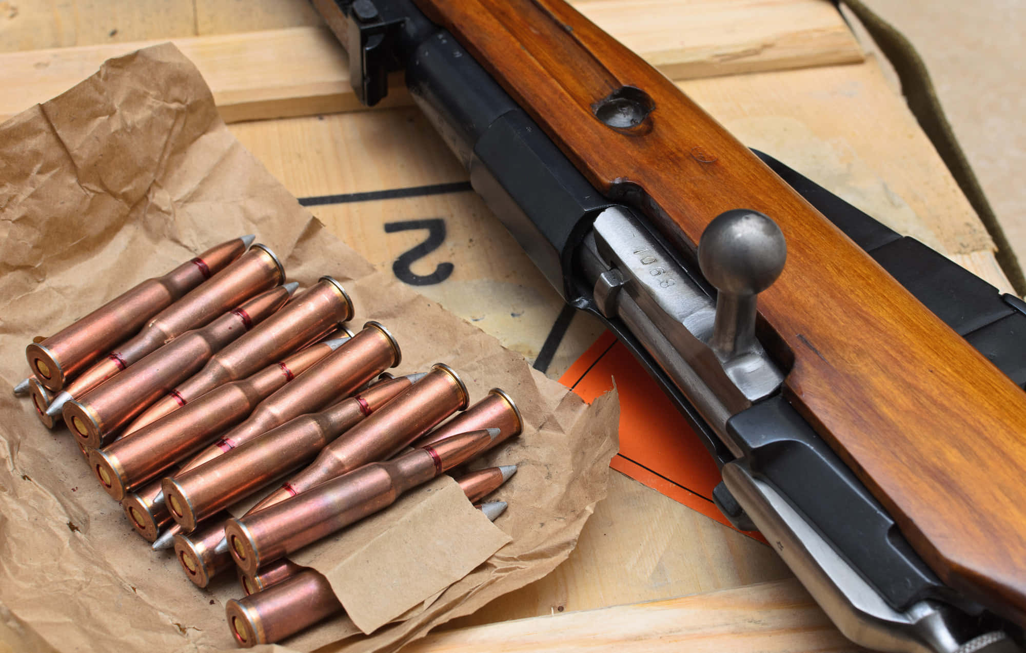 Rifle 桌布 壁紙 — 一支復古栓動步槍搭配木質槍托，放在一堆銅殼子彈旁邊，子彈置於皺巴巴的棕色紙上。