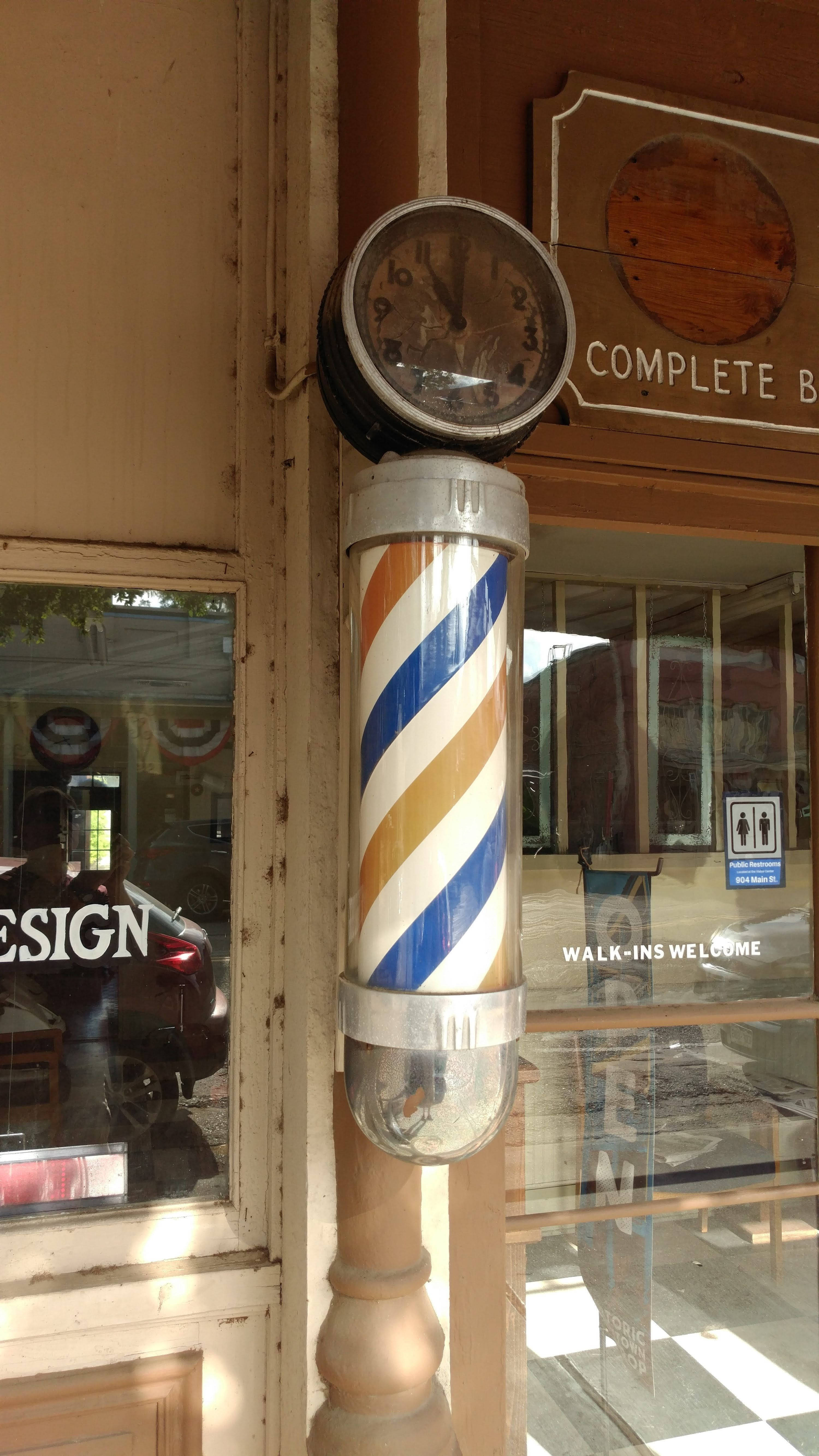 Barber 桌布 壁紙 — 一支復古理髮店旋轉燈柱，帶有藍、白、金色條紋，矗立在一間經典理髮店外，頂部有一個時鐘。