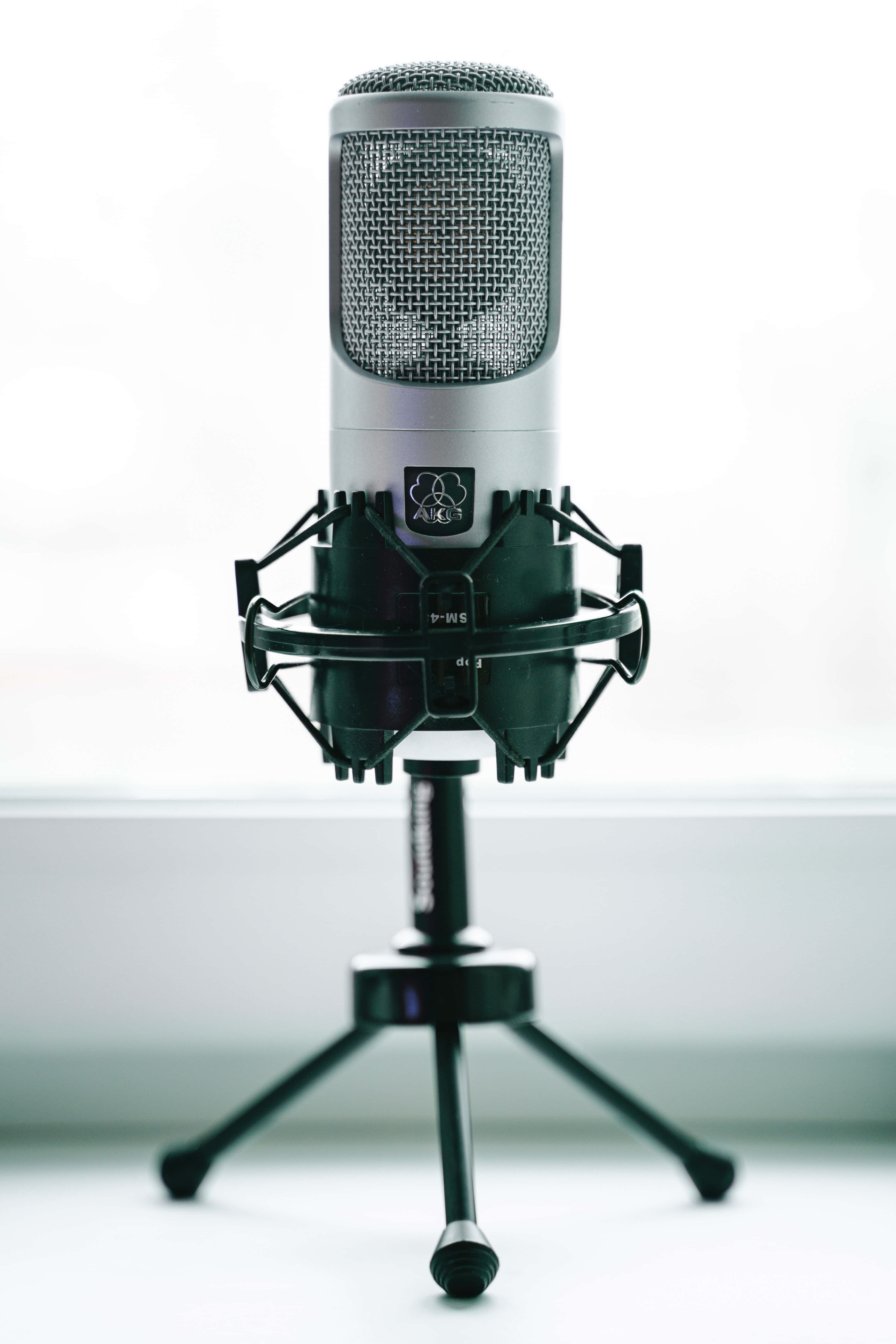 Microphone 桌布 壁紙 — 一支時尚的銀色電容麥克風，安裝在黑色防震架和三腳架上，背景是明亮的窗戶。