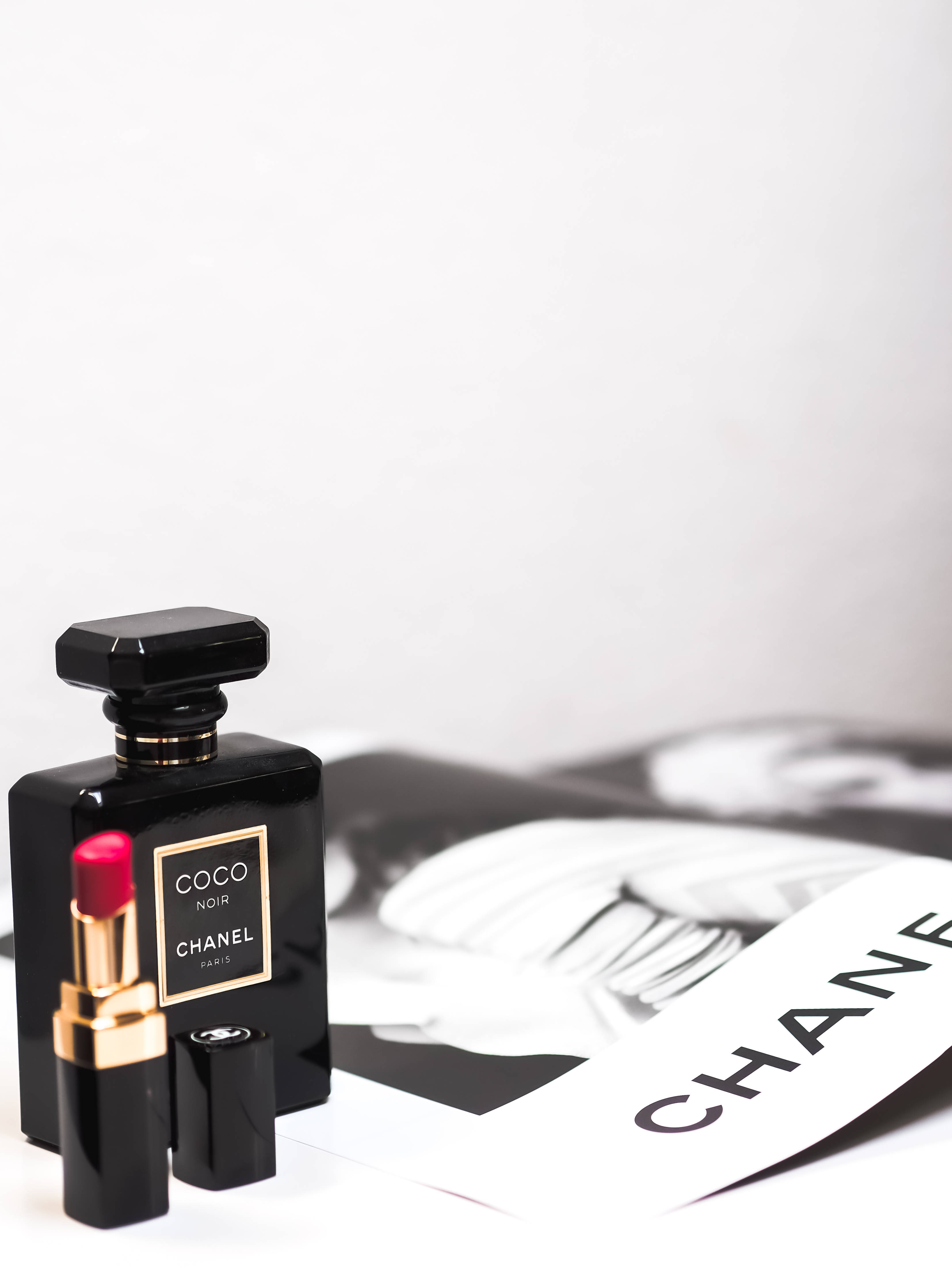Chanel 桌布 壁紙 — 一支時尚的黑色 Chanel Coco Noir 香水瓶和相配的口紅放在白色表面上，旁邊是一本 Chanel 雜誌內頁。