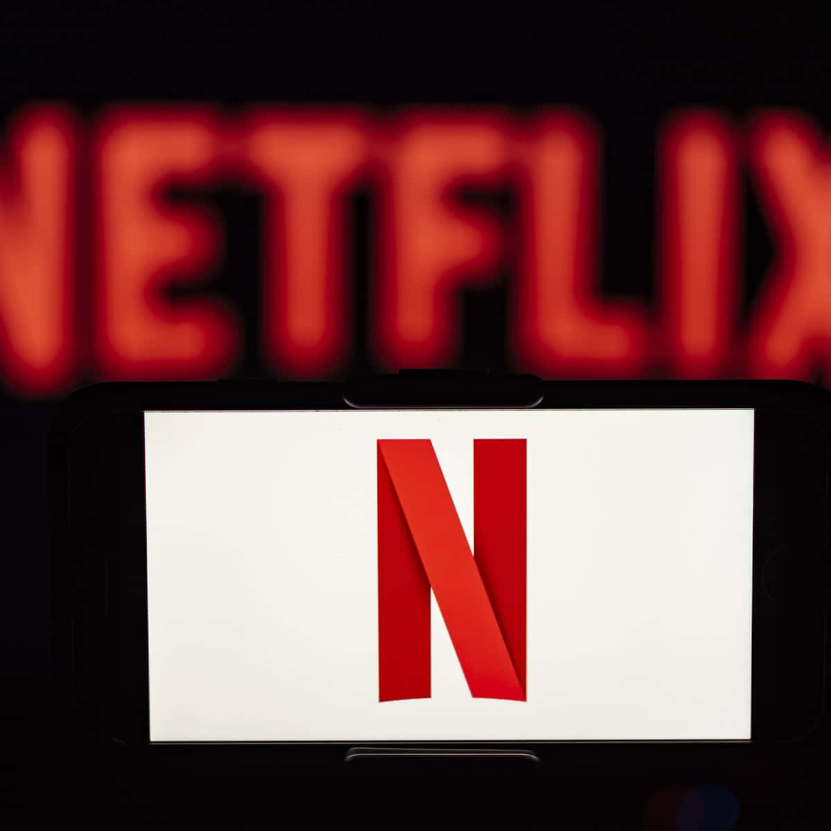 Netflix 桌布 壁紙 — 一支智慧型手機在深色背景上顯示紅色 Netflix 標誌，背後有模糊的紅色 Netflix 標牌。
