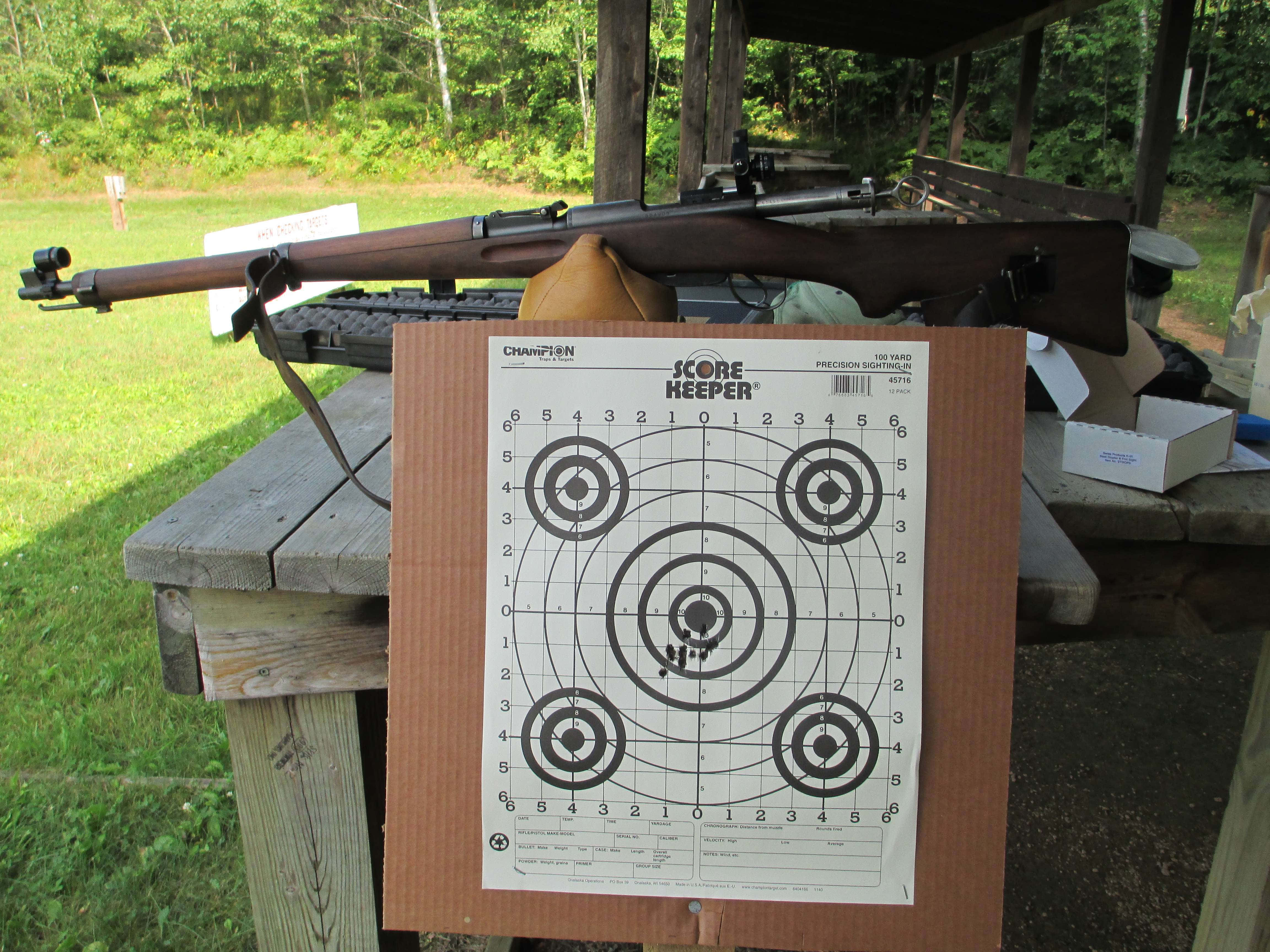 Rifle 桌布 壁紙 — 一支木製步槍擱在長凳上，旁邊放著一張靶紙，靶心附近有彈孔。