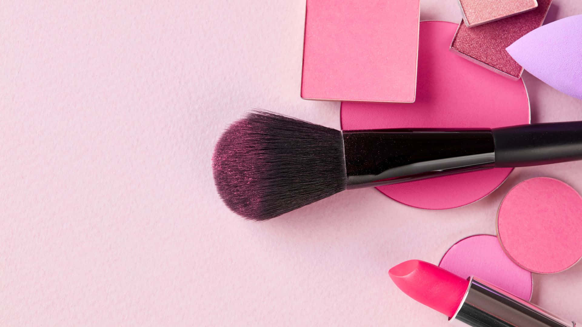 Pink Makeup 桌布 壁紙 — 一支柔軟的粉紅色化妝刷和唇膏放在散落的粉紅色腮紅粉餅上，表面是粉彩粉紅色紋理。