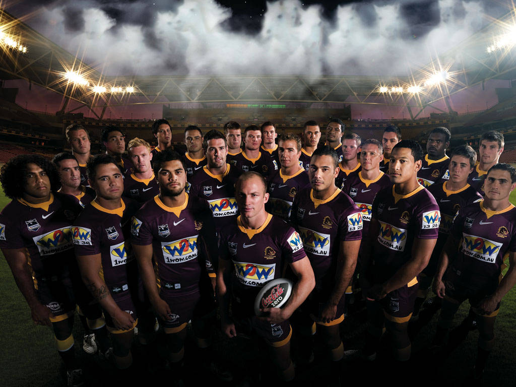 Nrl 桌布 壁紙 — 一支橄欖球聯賽隊伍身穿棗紅色和金色球衣，在煙霧繚繞的球場燈光下擺姿勢。