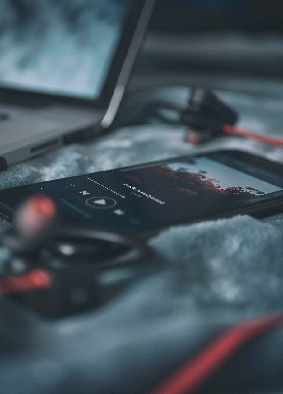 音樂 桌布 壁紙 — 一支正在播放音樂的智慧型手機放在柔軟的灰色毛毯上，旁邊有一台筆記型電腦和紅色耳塞，營造出舒適的室內環境。