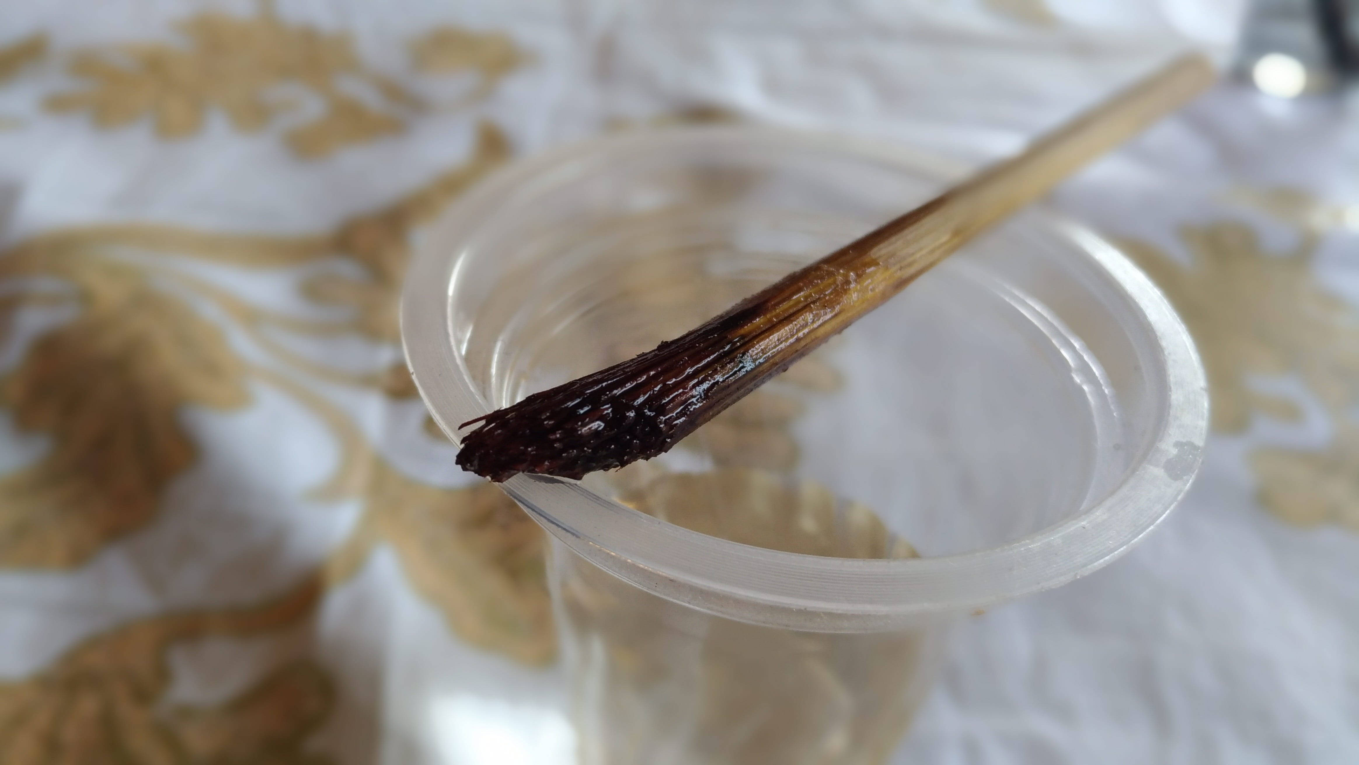 Painting 桌布 壁紙 — 一支深棕色刷毛的畫筆放在透明塑膠杯中，置於帶金色花卉圖案的白色布料上。