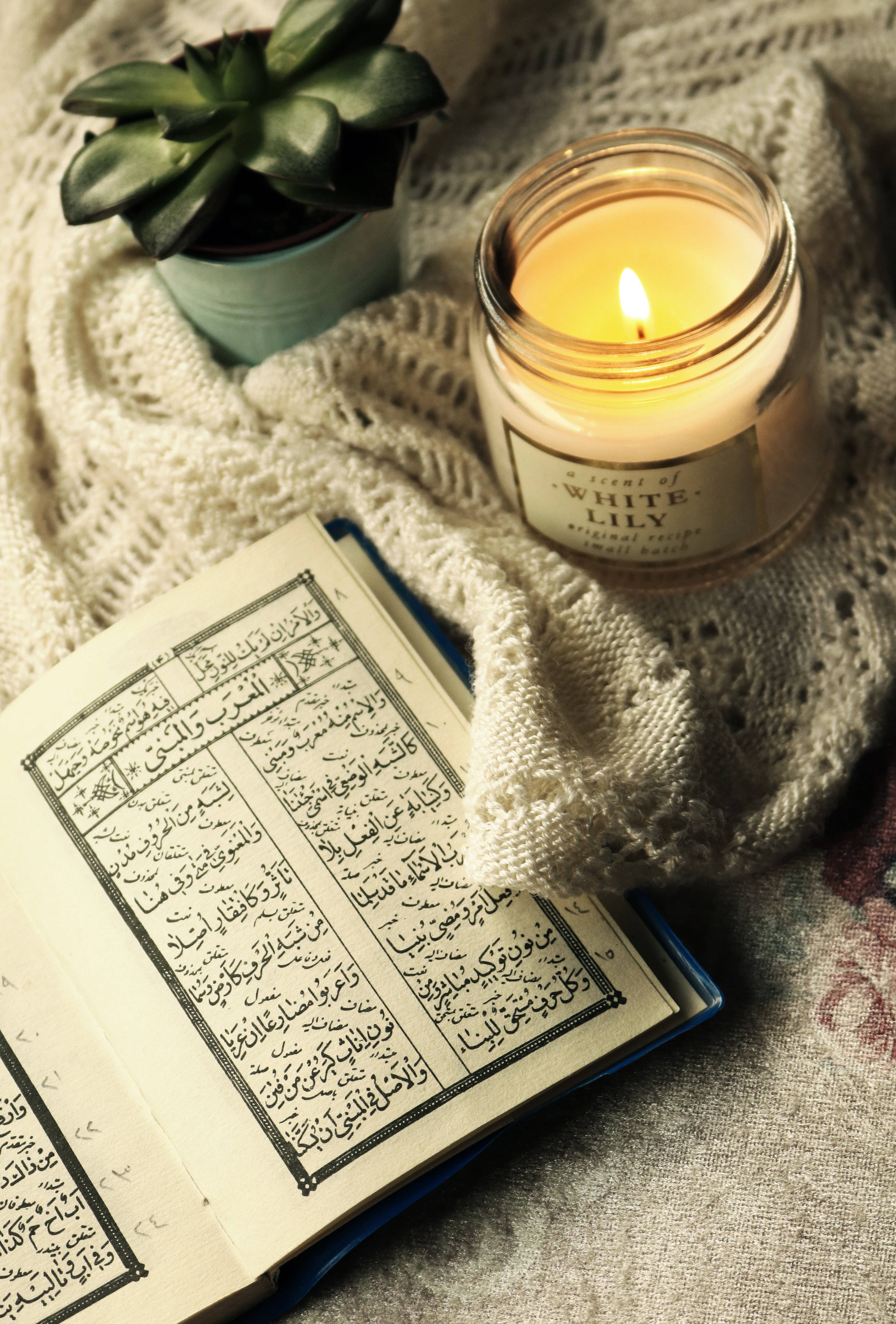 Best Islamic 桌布 壁紙 — 一支溫暖的蠟燭旁邊放著一本打開的阿拉伯文書本，擱在舒適的針織毛毯上，附近還有一棵小多肉植物。