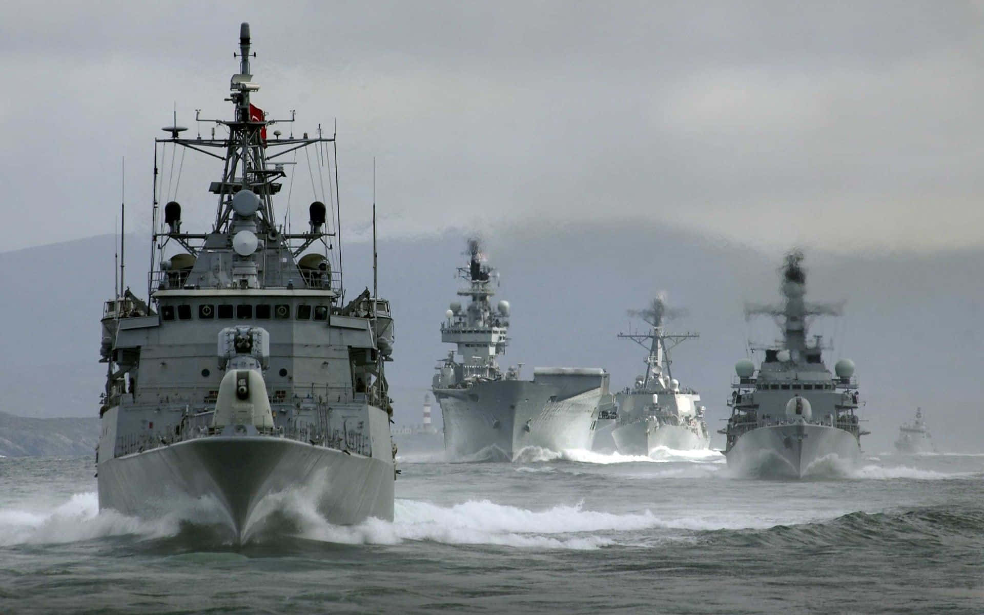 Navy 桌布 壁紙 — 一支灰色軍艦艦隊在多雲的天空下，在遠處群山的背景下，破浪前行。
