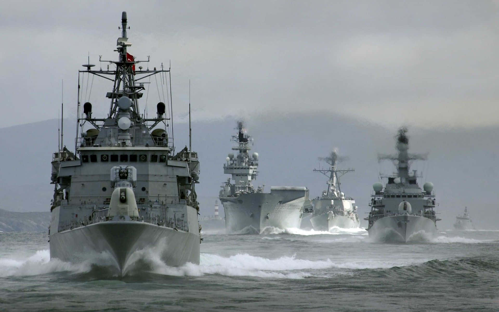 Navy 桌布 壁紙 — 一支灰色軍艦艦隊在多雲的天空下，背景有遠處的山脈，穿越波濤洶湧的水域。